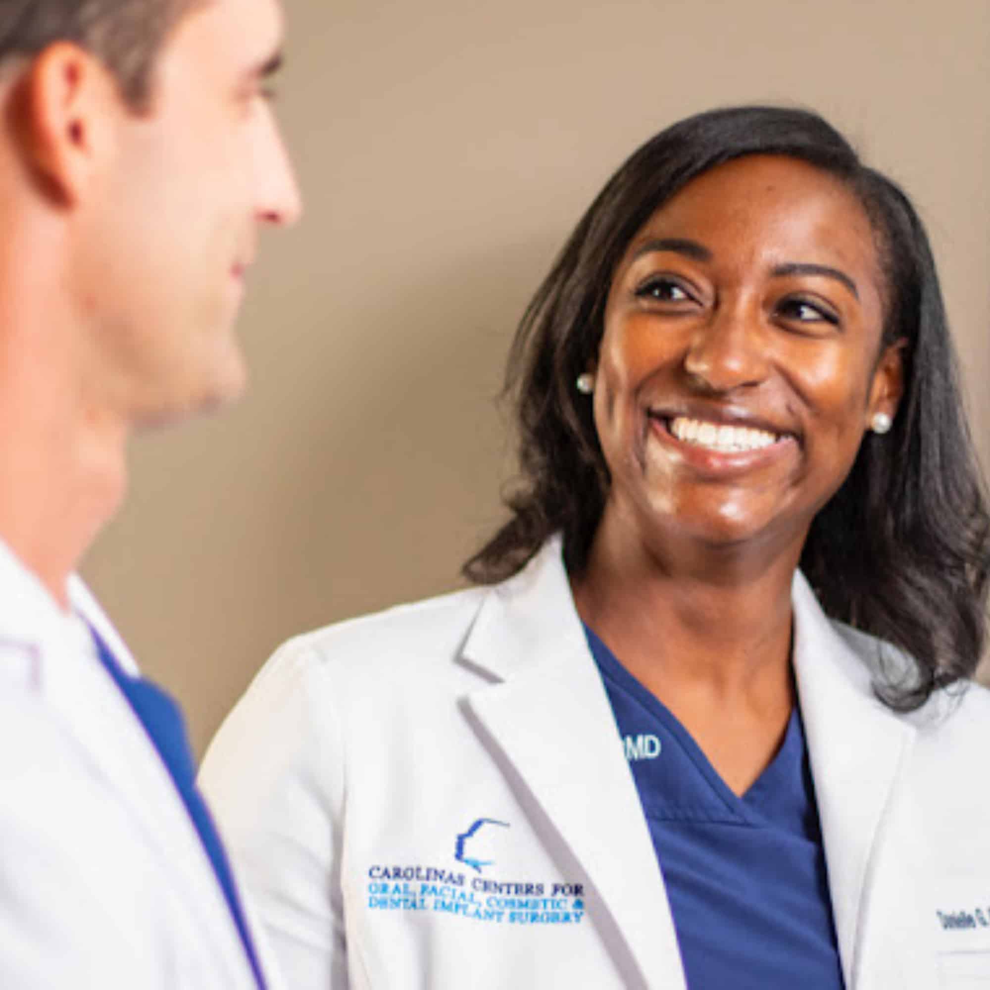 Two people in white coats smiling and conversing. Text indicates "Carolina Centers for Oral, Facial, Cosmetic & Dental Implant Surgery." Neutral background.