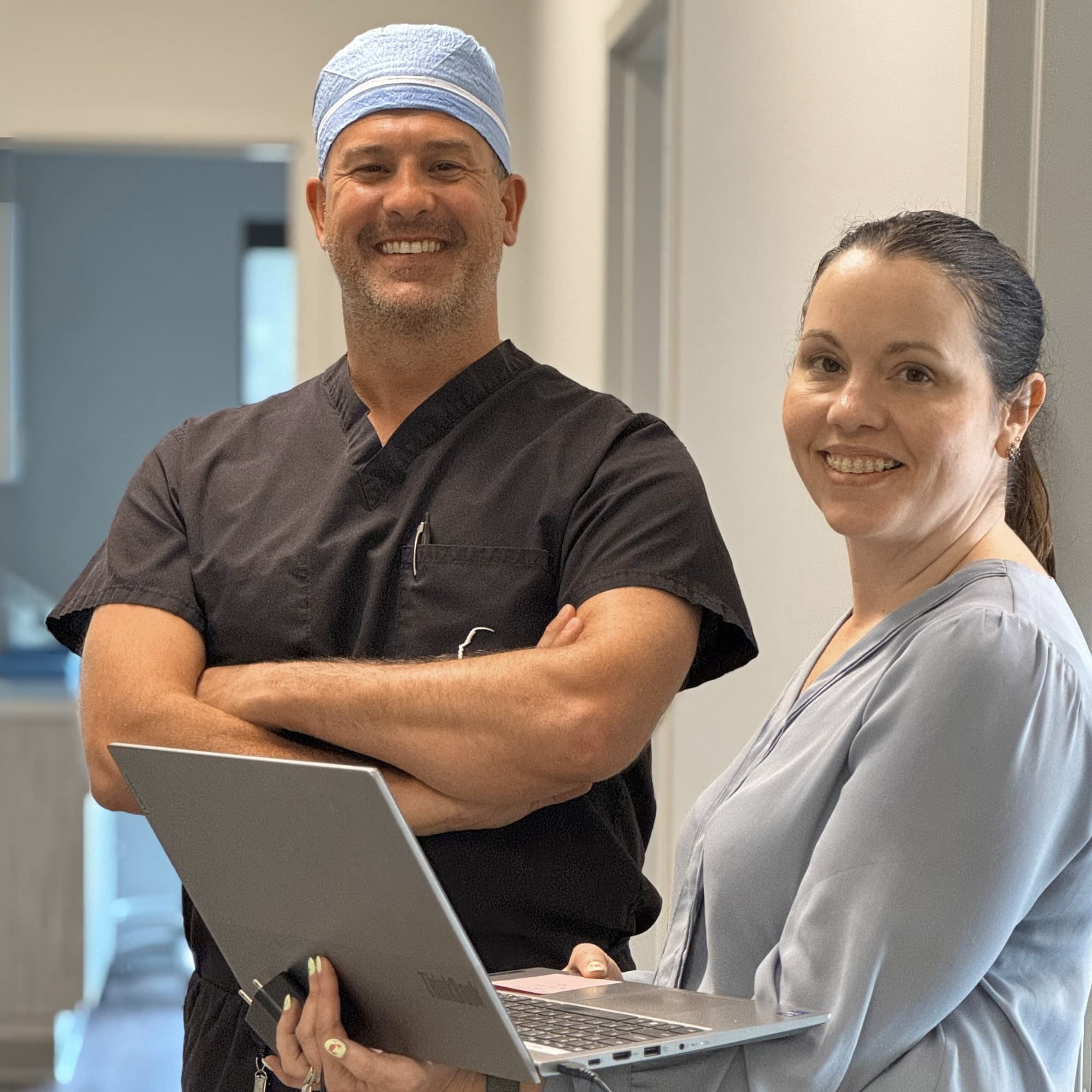 Two smiling people in medical attire stand in a hallway, one holding a laptop and the other wearing a blue surgical cap.
