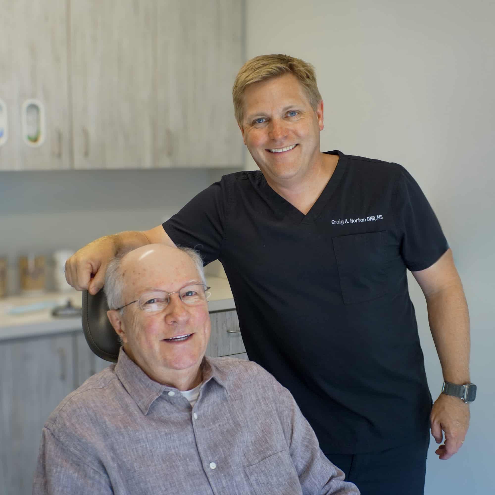 Two people in a dental office, one seated and one standing, smiling at the camera, with cabinets in the background.
