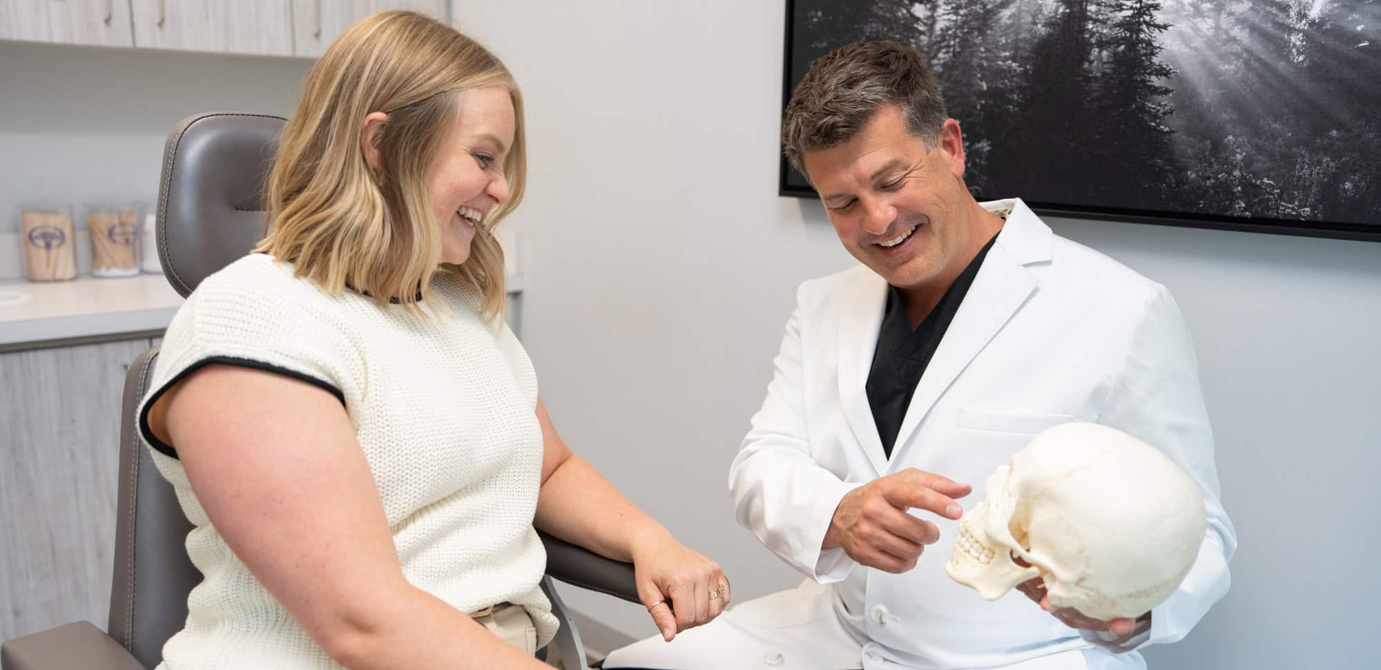 Two people in a medical office, one showing a skull model. They appear engaged and smiling, seated near cabinetry and wall art.