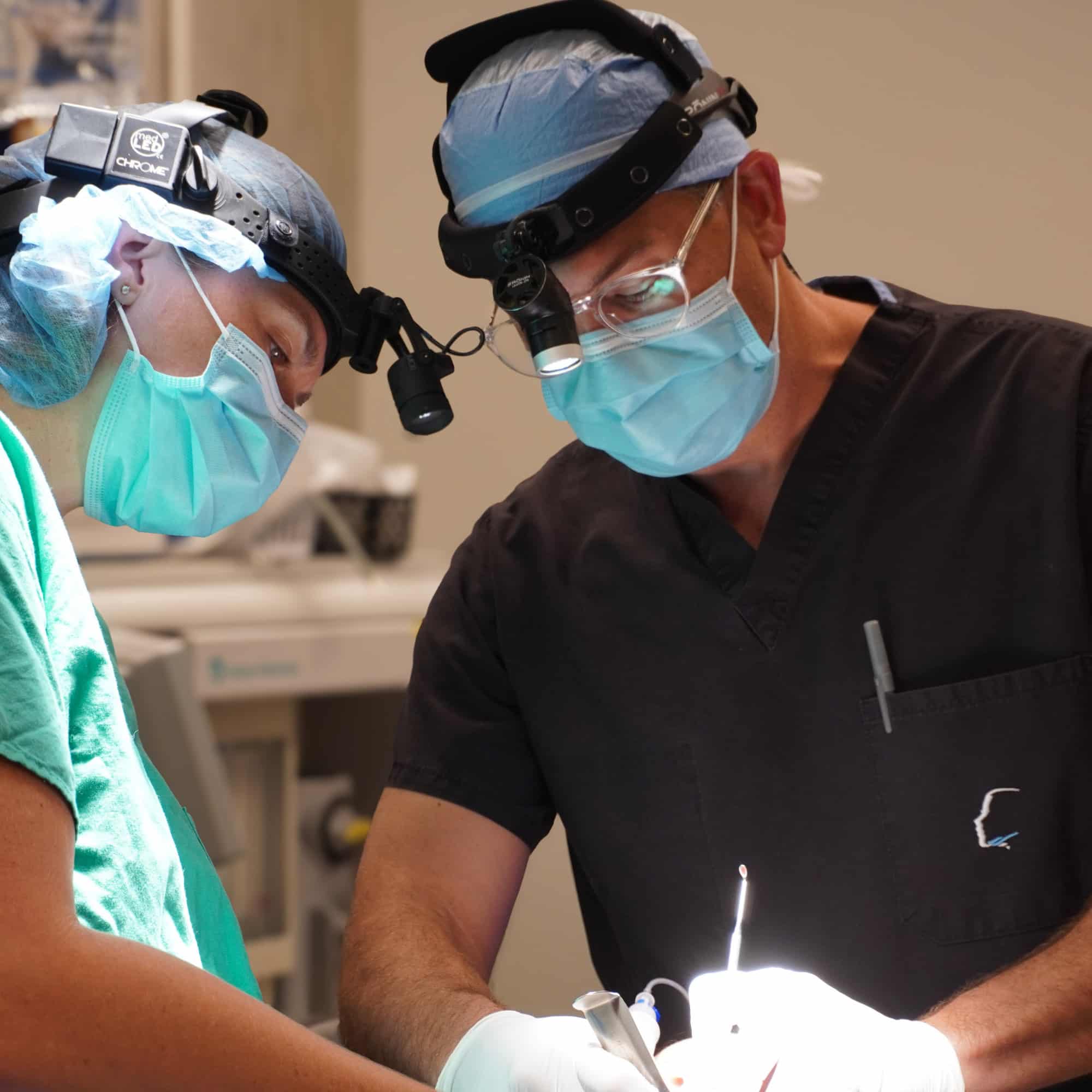 Two people in surgical attire focus intently during a procedure, using headlamps and surgical tools in a well-lit operating room.