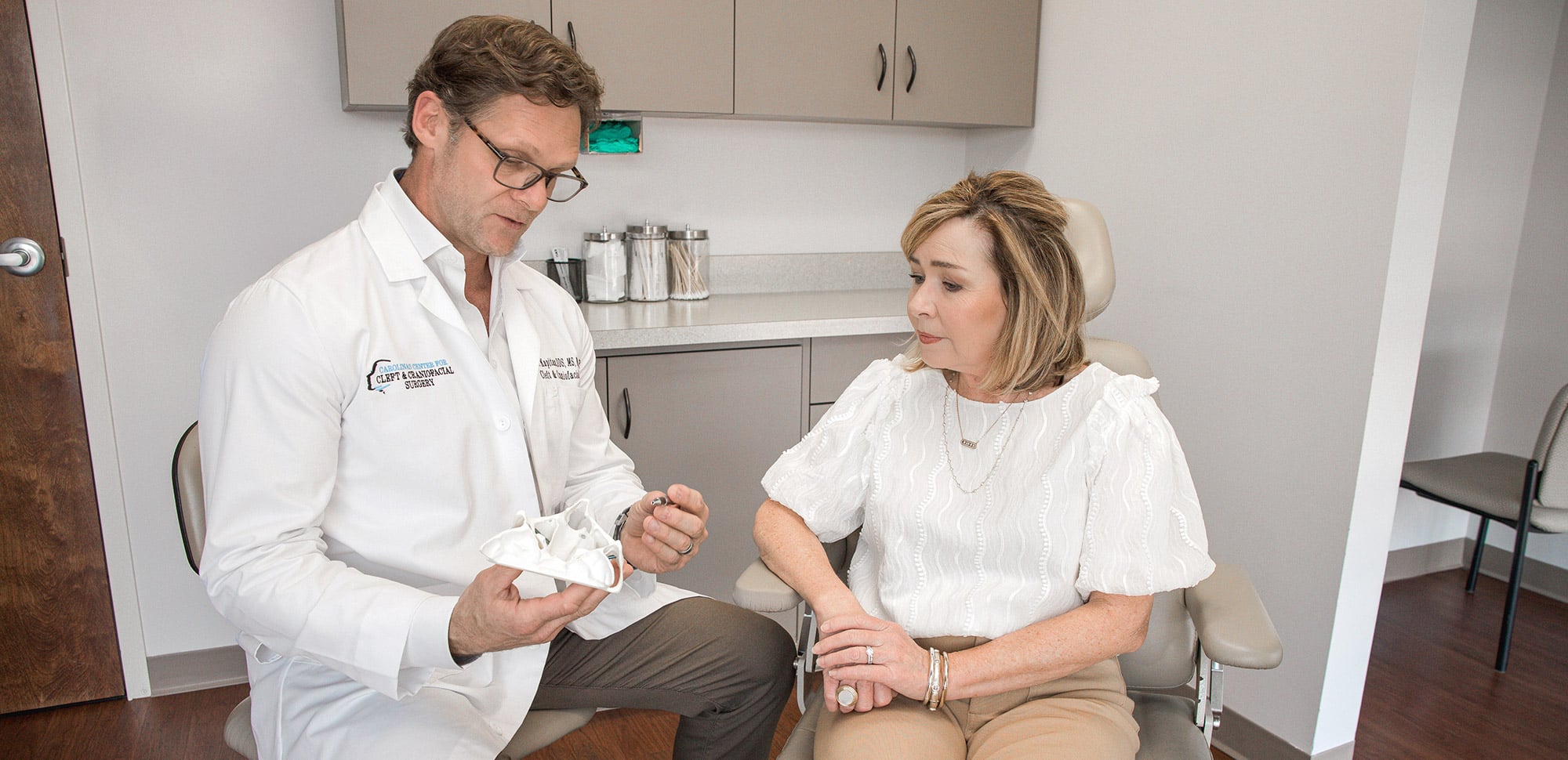 Two people in a medical office, one in a white coat discussing a dental model. Cabinets and supplies are in the background.