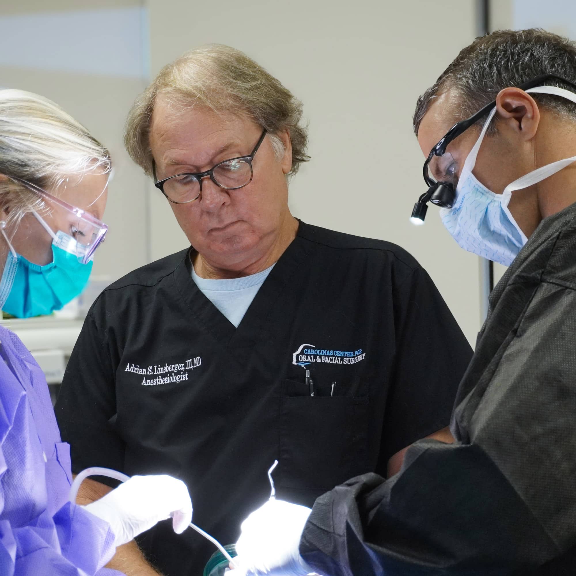 Three persons in medical attire focus on a task. One wears a magnifying headlamp, suggesting a medical procedure or training in a clinical setting.