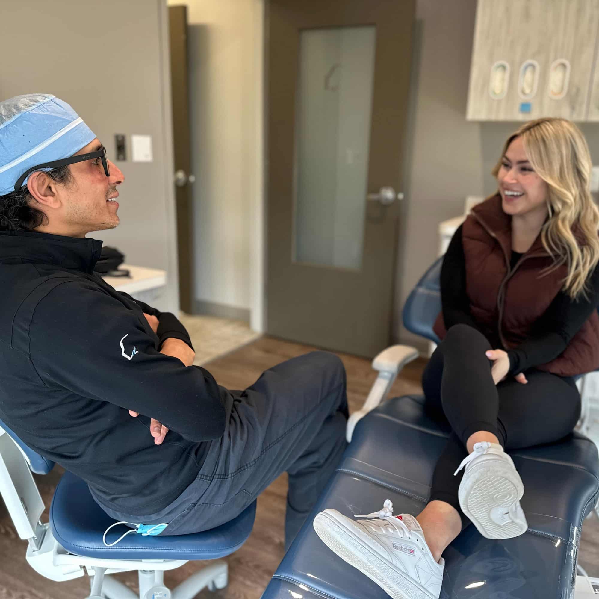 Two people are in a dental office, sitting on chairs and smiling at each other, engaging in a conversation.