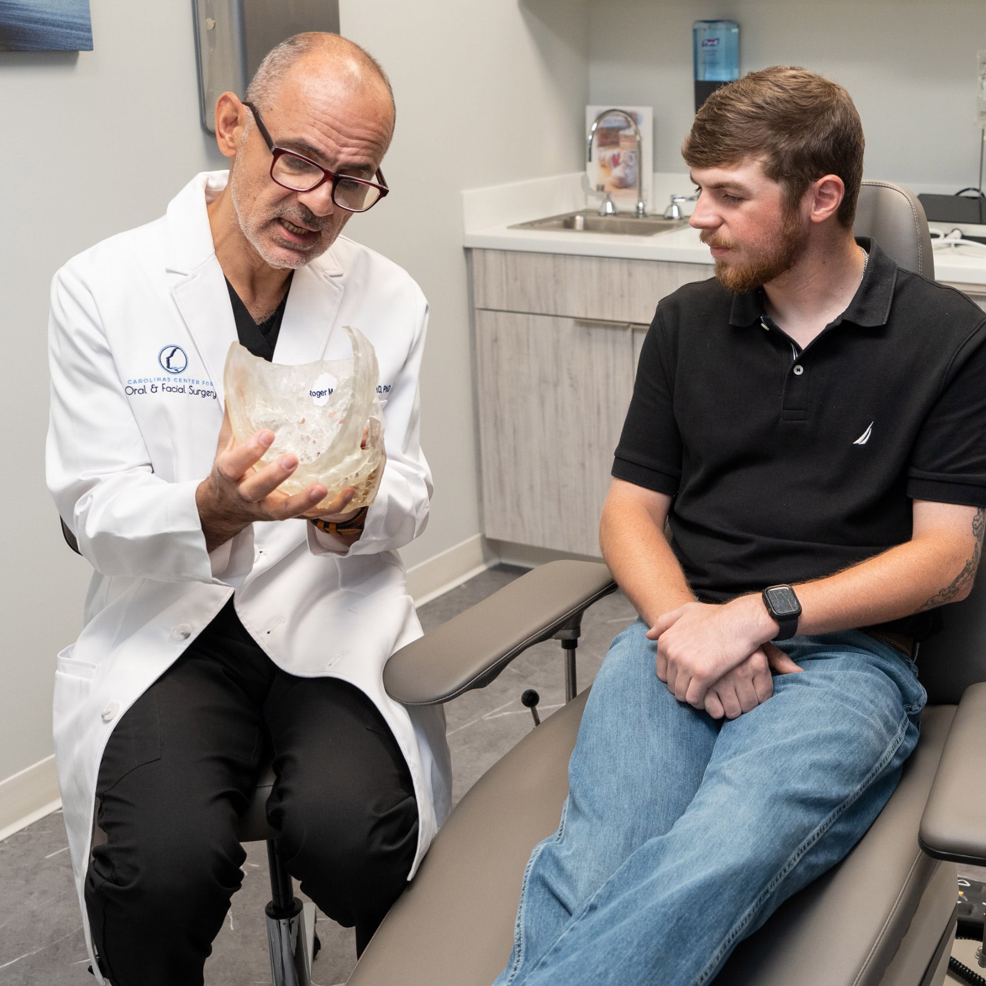 Two people in a medical office, one holding a transparent skull model, likely discussing a medical procedure.