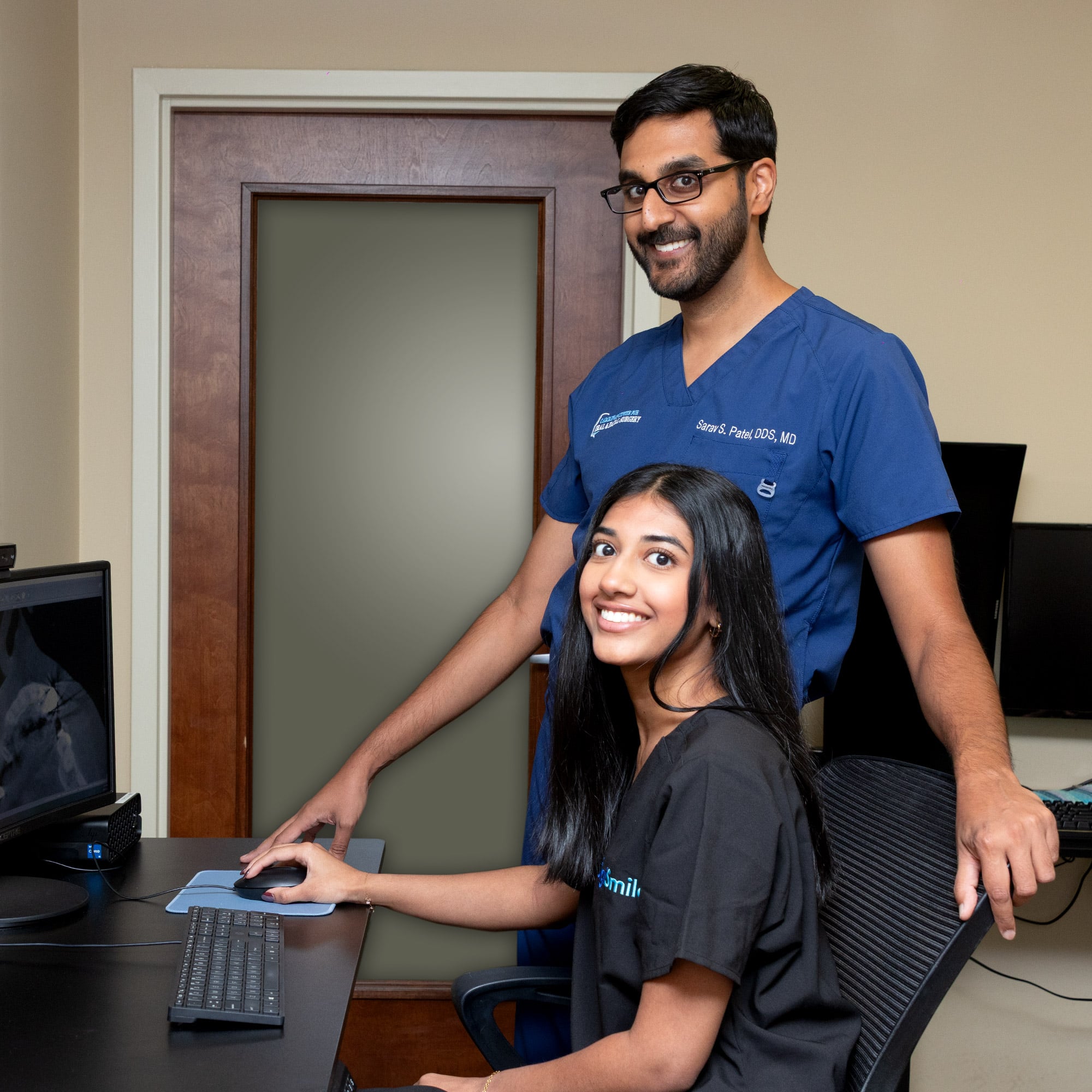 Two people are smiling in an office, with one seated at a computer and the other standing beside them in medical scrubs.