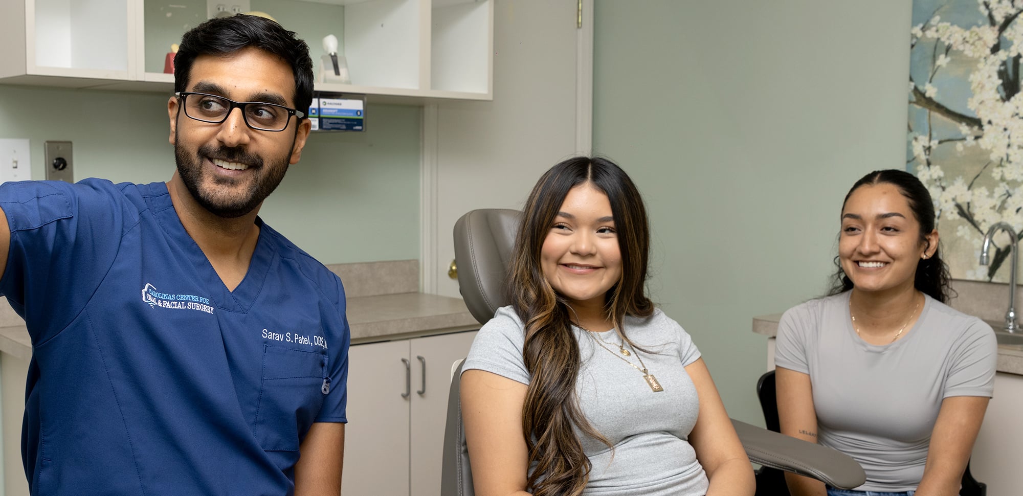 A person in dental scrubs gestures smiling, sitting with two other smiling individuals in a clinic setting, against a floral wall painting.