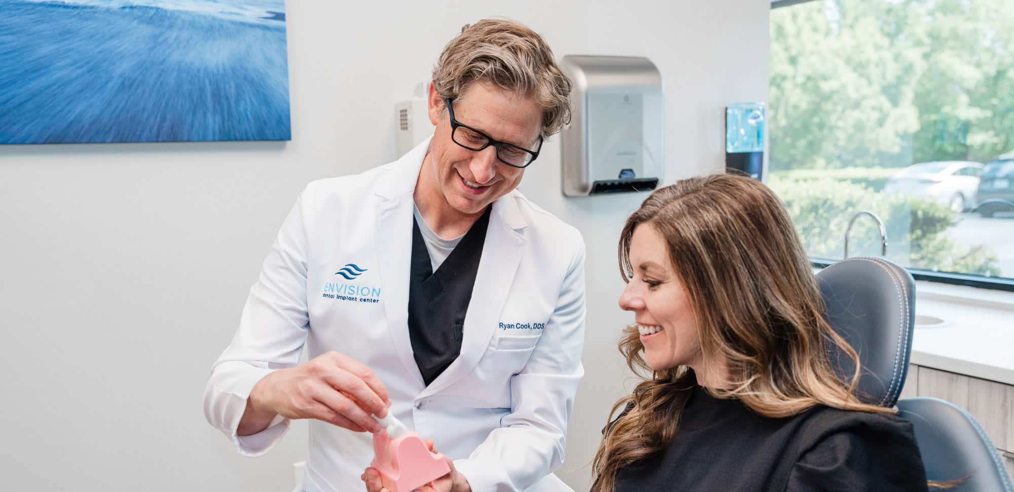 Two persons in a dental clinic, one demonstrating dental care with a model. They are smiling, seated near a large window.