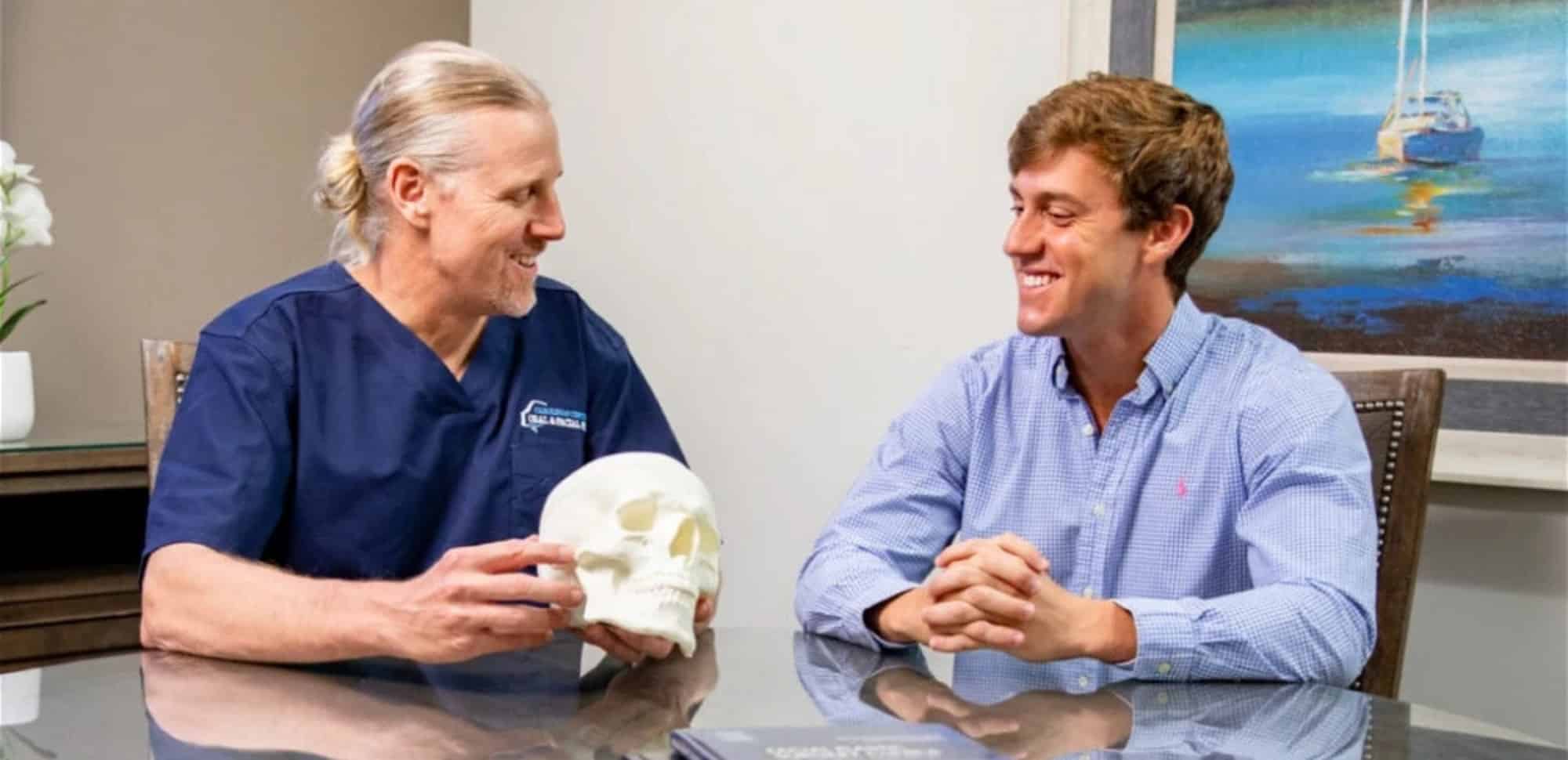 Two persons seated at a table, engaged in discussion. One holds a skull model; a painting of a sailboat is in the background.