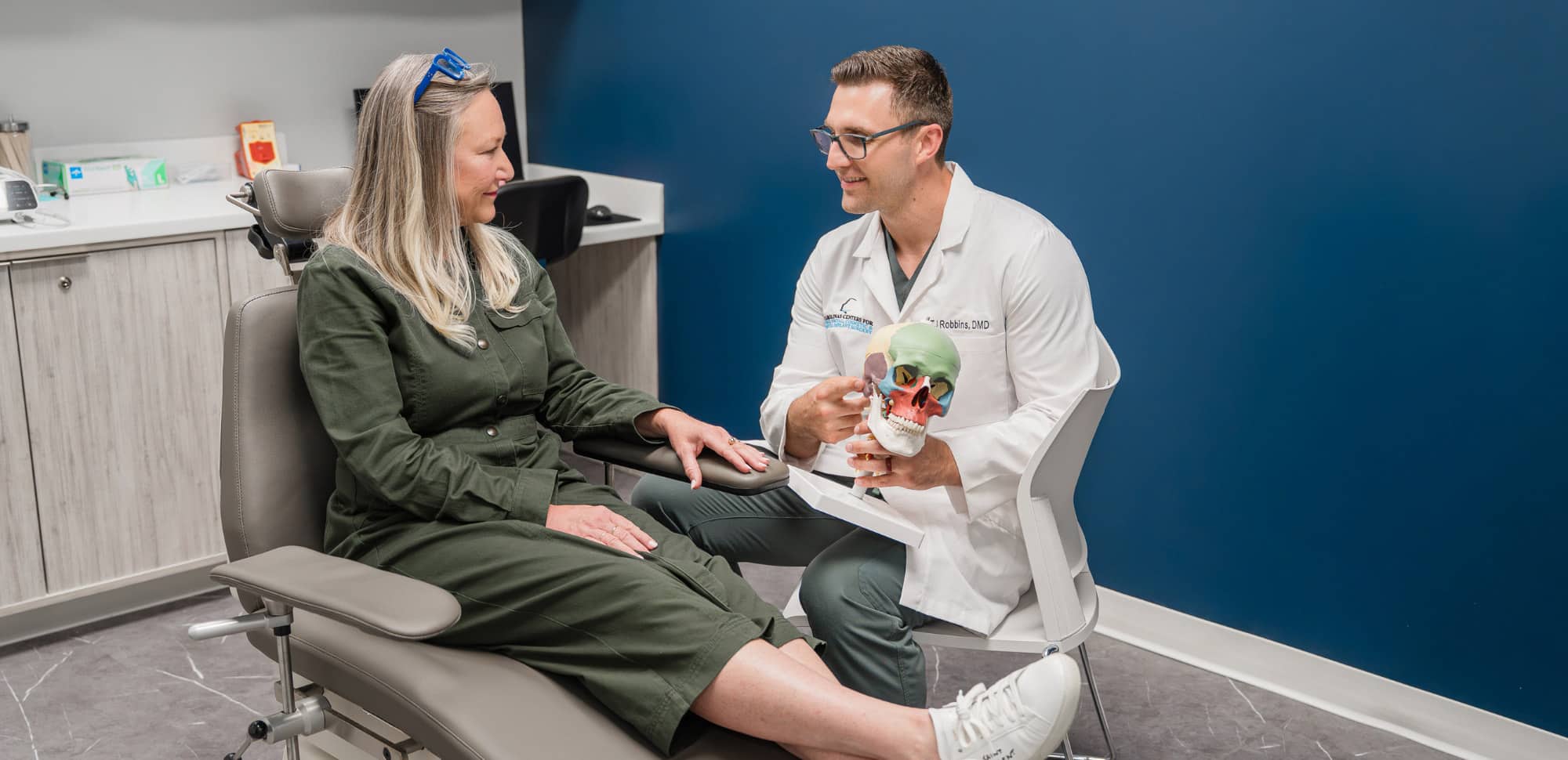 A person in a lab coat discusses with seated person, holding a colorful skull model in a modern office setting.