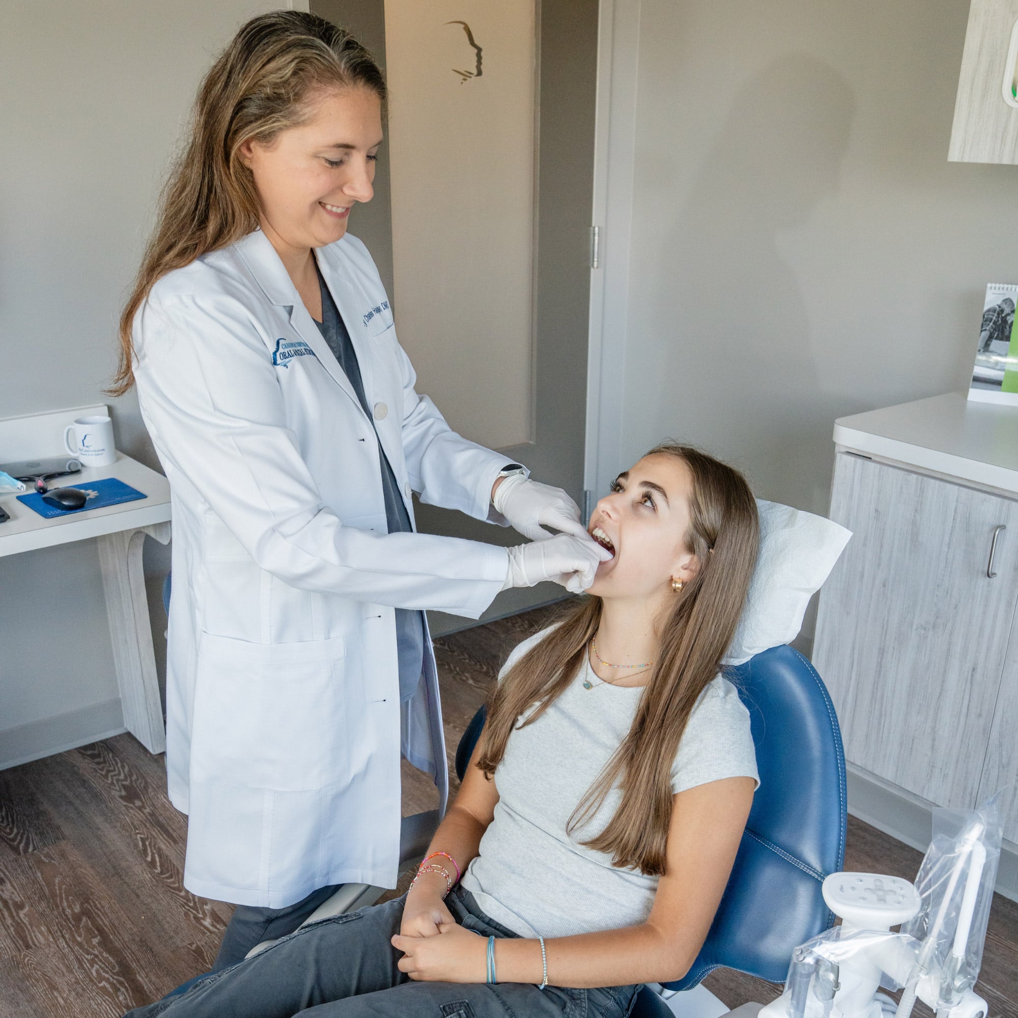 A person in a white coat examines another person's teeth in a dental office, with medical tools nearby on a table.