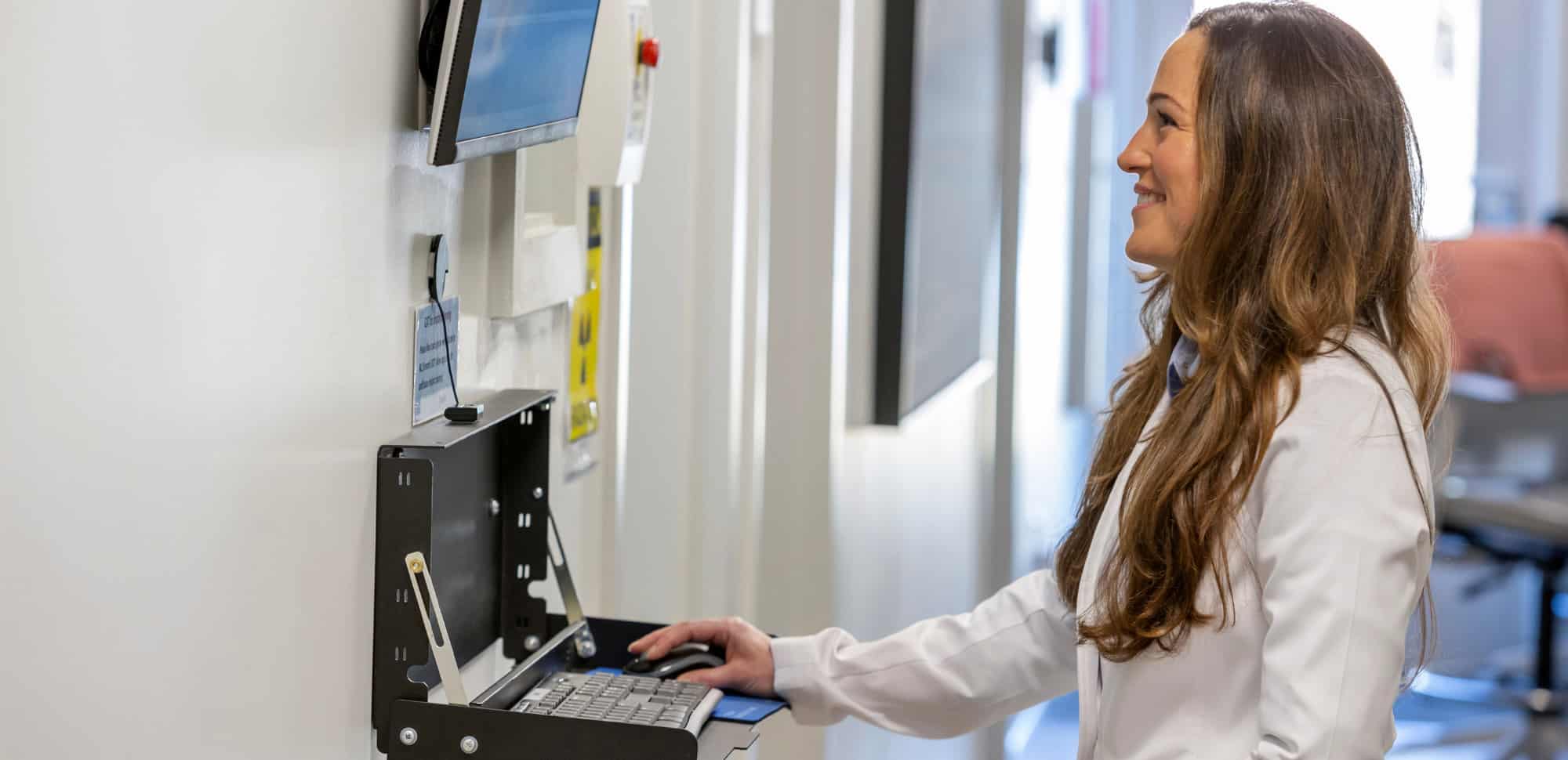 A person in a white coat operates a computer mounted on a wall in a clinical setting, smiling while working.