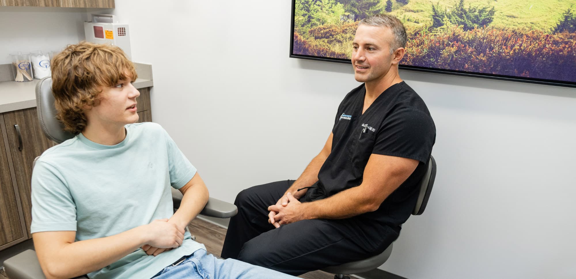 A person in scrubs talks with a seated person in a casual setting. The room has a nature photograph on the wall.