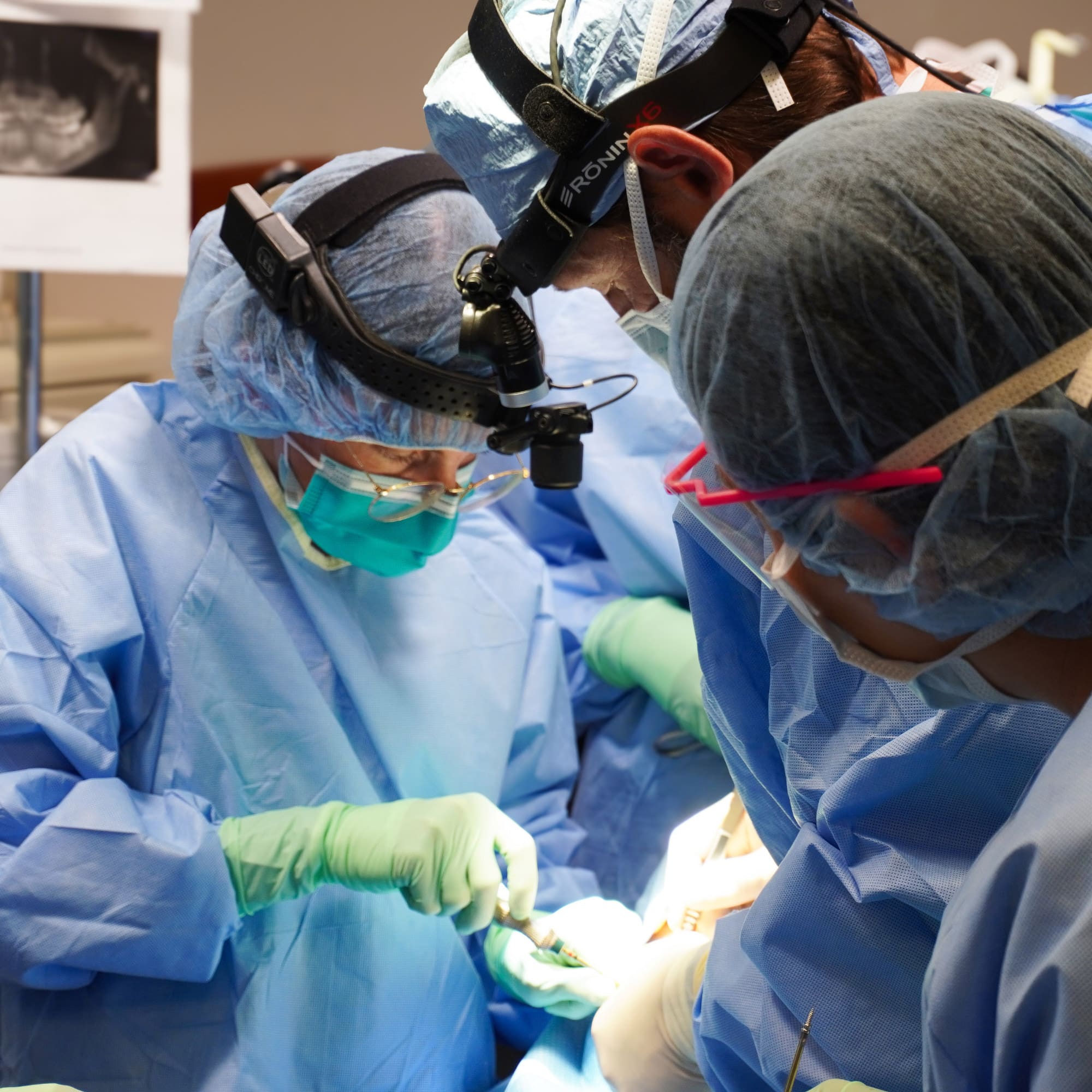Three people in surgical attire perform an operation, wearing masks and headlamps, focused on a patient. Medical equipment and an X-ray are visible.