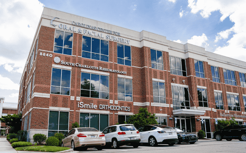 A brick building houses various medical offices, including Carolinas Center for Oral Surgery and iSmile Orthodontics. Several cars are parked outside.