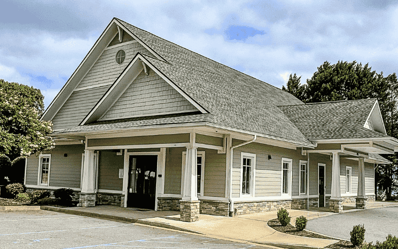 A modern, single-story building with a gabled roof and columns, surrounded by trees and shrubs, under a partly cloudy sky.
