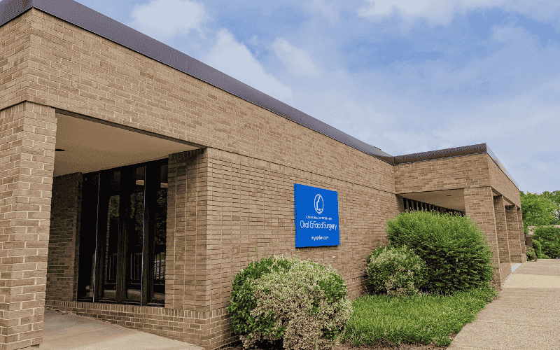 A brick building with a blue sign for an oral surgery center, surrounded by shrubs, under a partly cloudy sky.