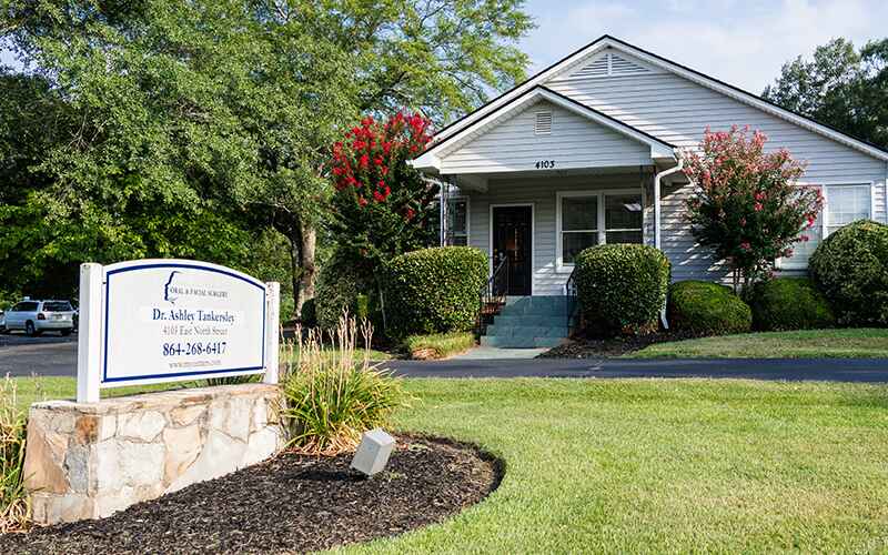 A single-story house with a porch is surrounded by greenery. A sign for "Dr. Ashley Tuckersley" stands in the manicured lawn.