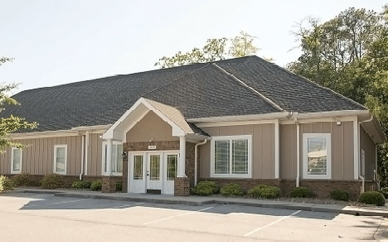 A beige single-story building with white trim and a dark roof. The entrance has double doors. Surrounding green foliage is visible.