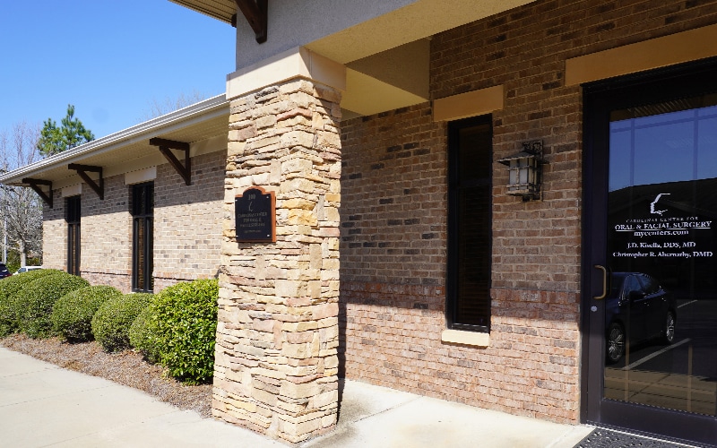 A brick and stone building with large windows and a glass entry door, surrounded by neatly trimmed shrubs, under a clear blue sky.