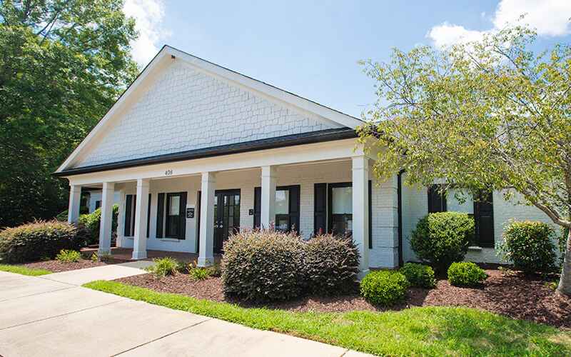 A small, white brick building with a gabled roof, columns, and manicured landscaping under a clear blue sky. No people are visible.