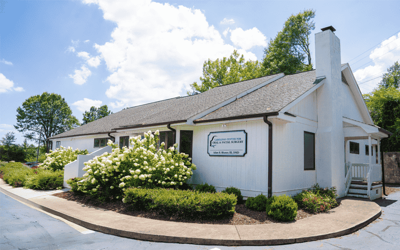 A white building with a chimney houses a dental surgery center, surrounded by lush shrubs and trees, under a bright, partly cloudy sky.