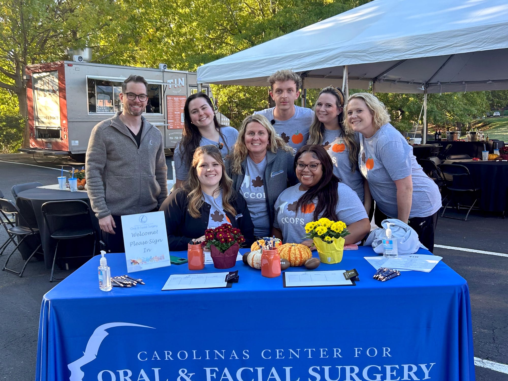 A group of people at an outdoor event table with fall decorations and a food truck in the background under a tent.