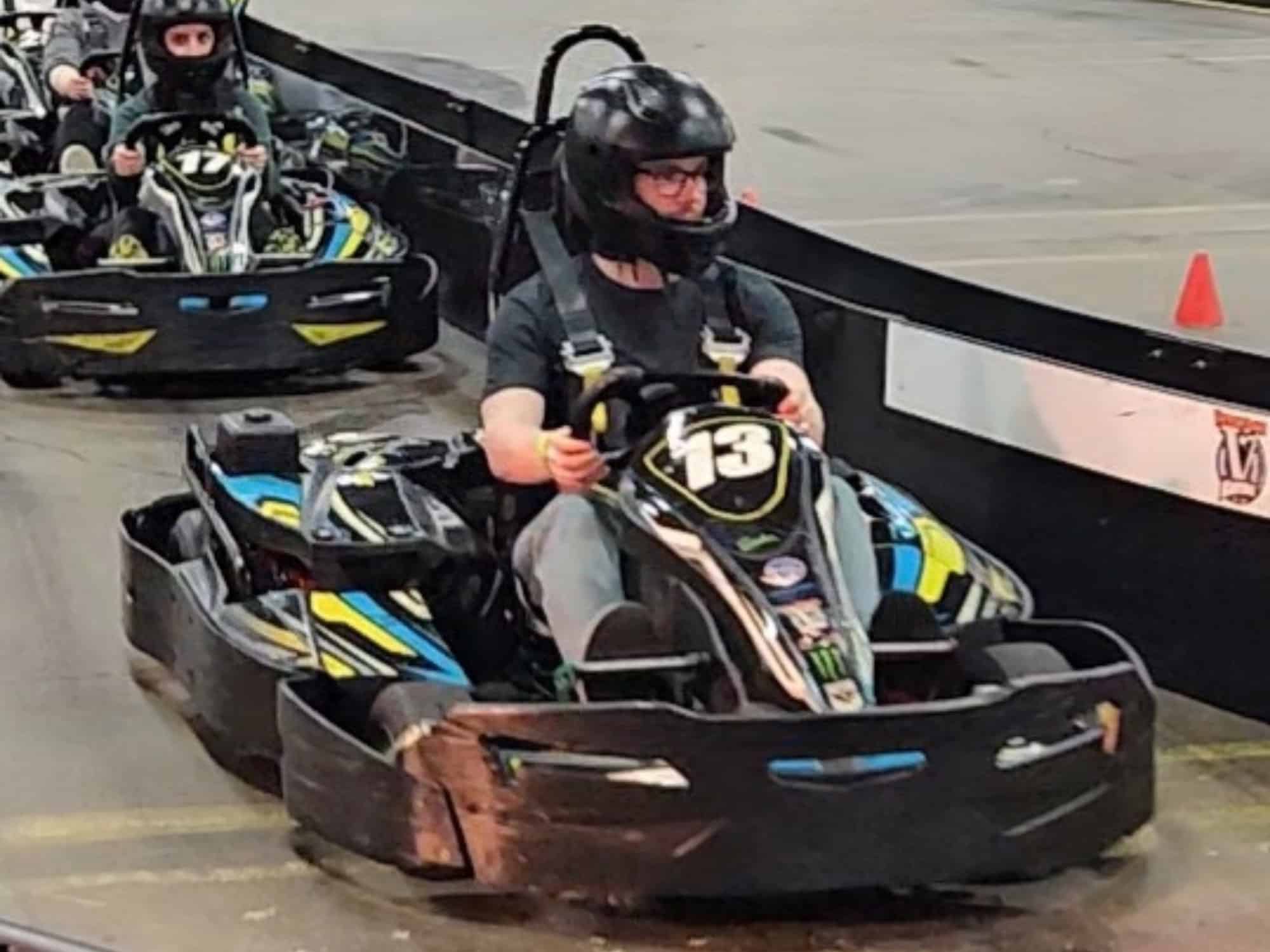 People driving go-karts indoors, wearing helmets. Focus on the leading kart number 13. Track barriers and cones visible in the background.