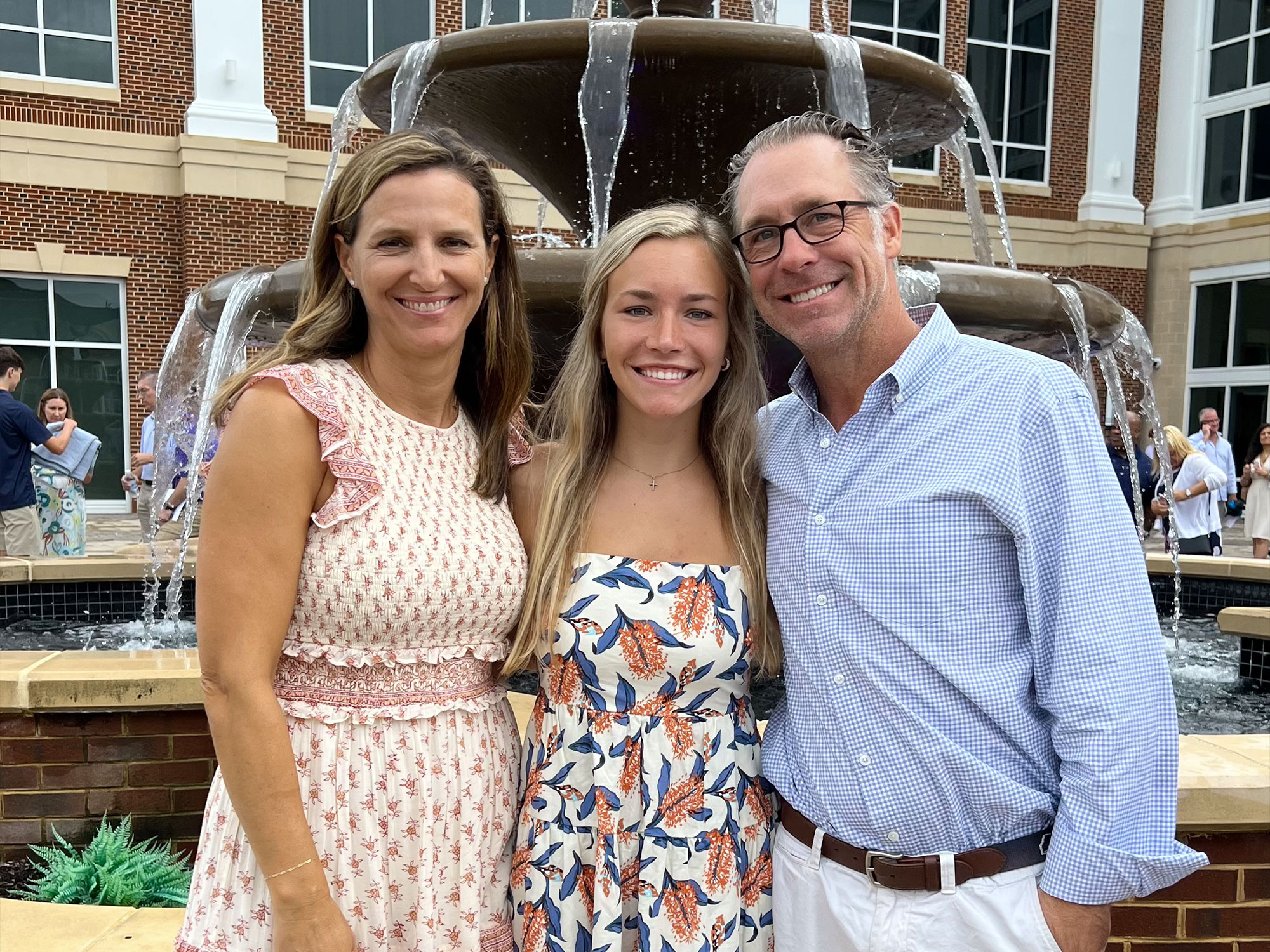 Three people smiling in front of a tiered fountain, with a brick building in the background. Social gathering in progress.