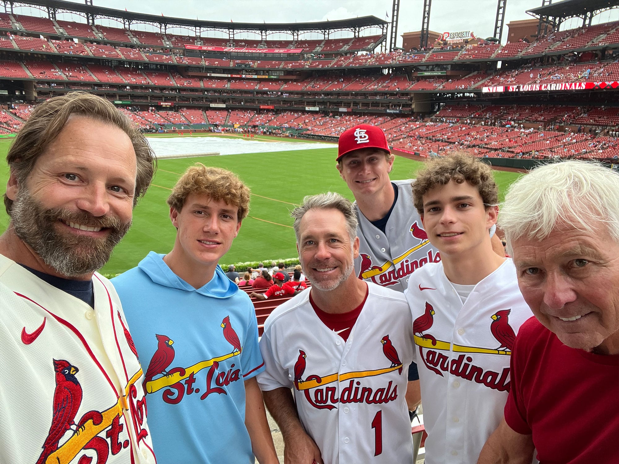 Five people wearing St. Louis Cardinals apparel pose for a photo at Busch Stadium, with the baseball field visible in the background.