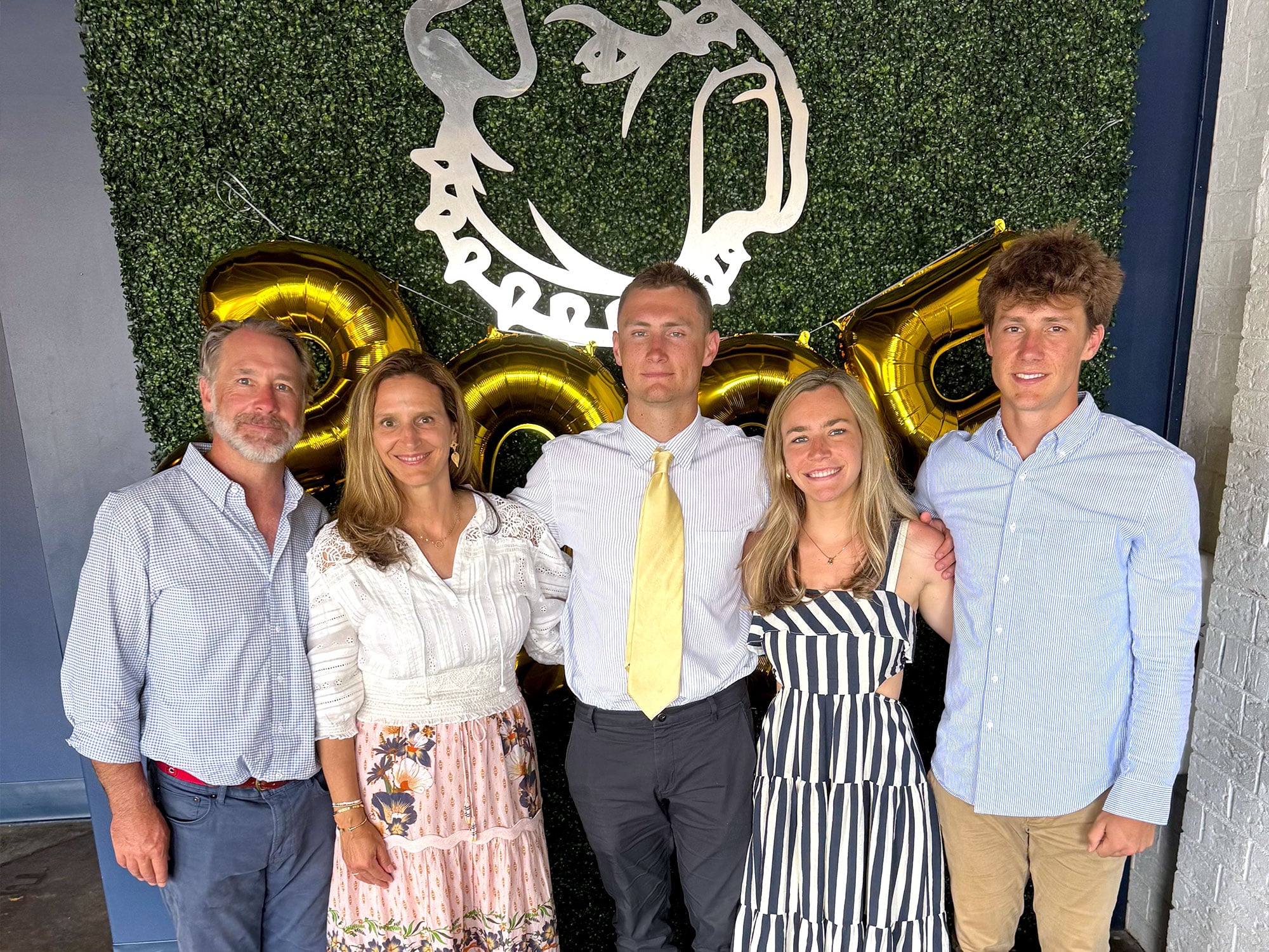 Five people stand together, smiling in front of gold balloons and greenery, featuring a stylized emblem above. Casual, celebratory atmosphere.