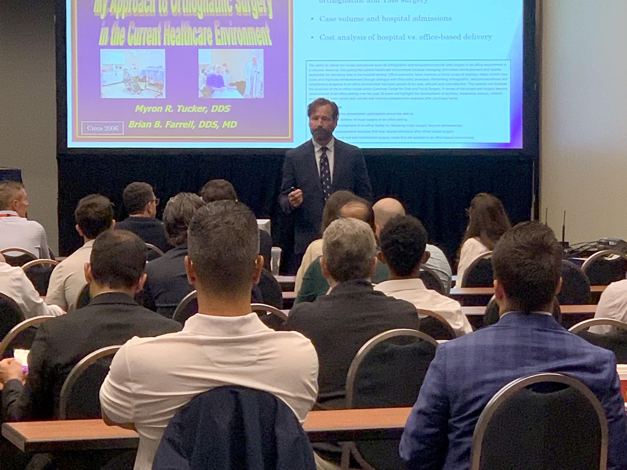 A person presents on orthognathic surgery at a conference. Attendees sit and listen, with slides projected in front of them.