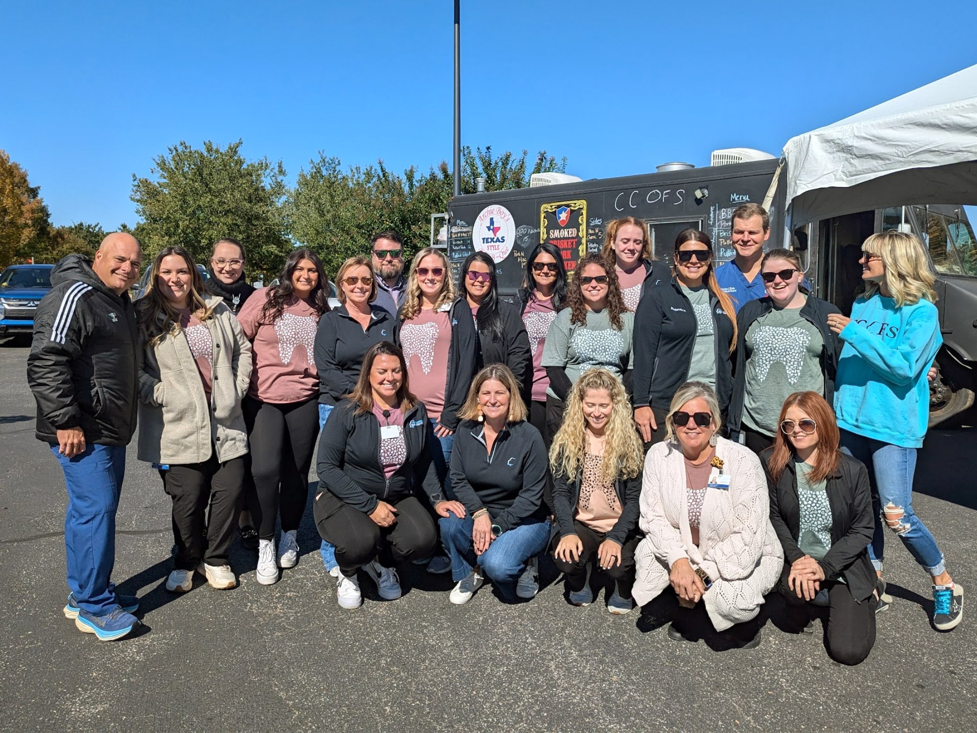 A group of people smiling outside near a food truck under a clear blue sky, surrounded by trees.