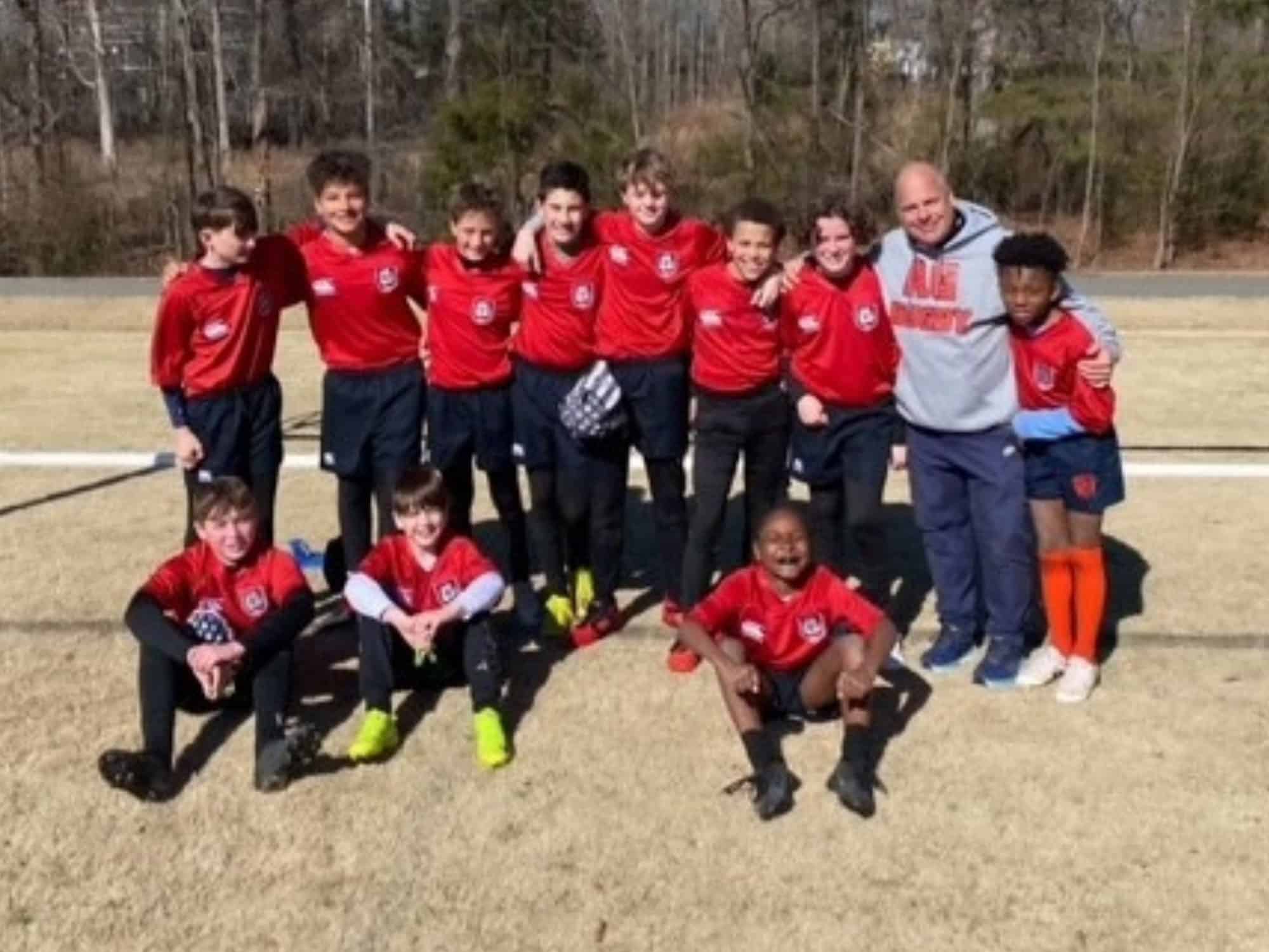 A soccer team smiles outdoors, wearing red jerseys. Eleven kids gather with one person. Trees and grass are visible in the background.