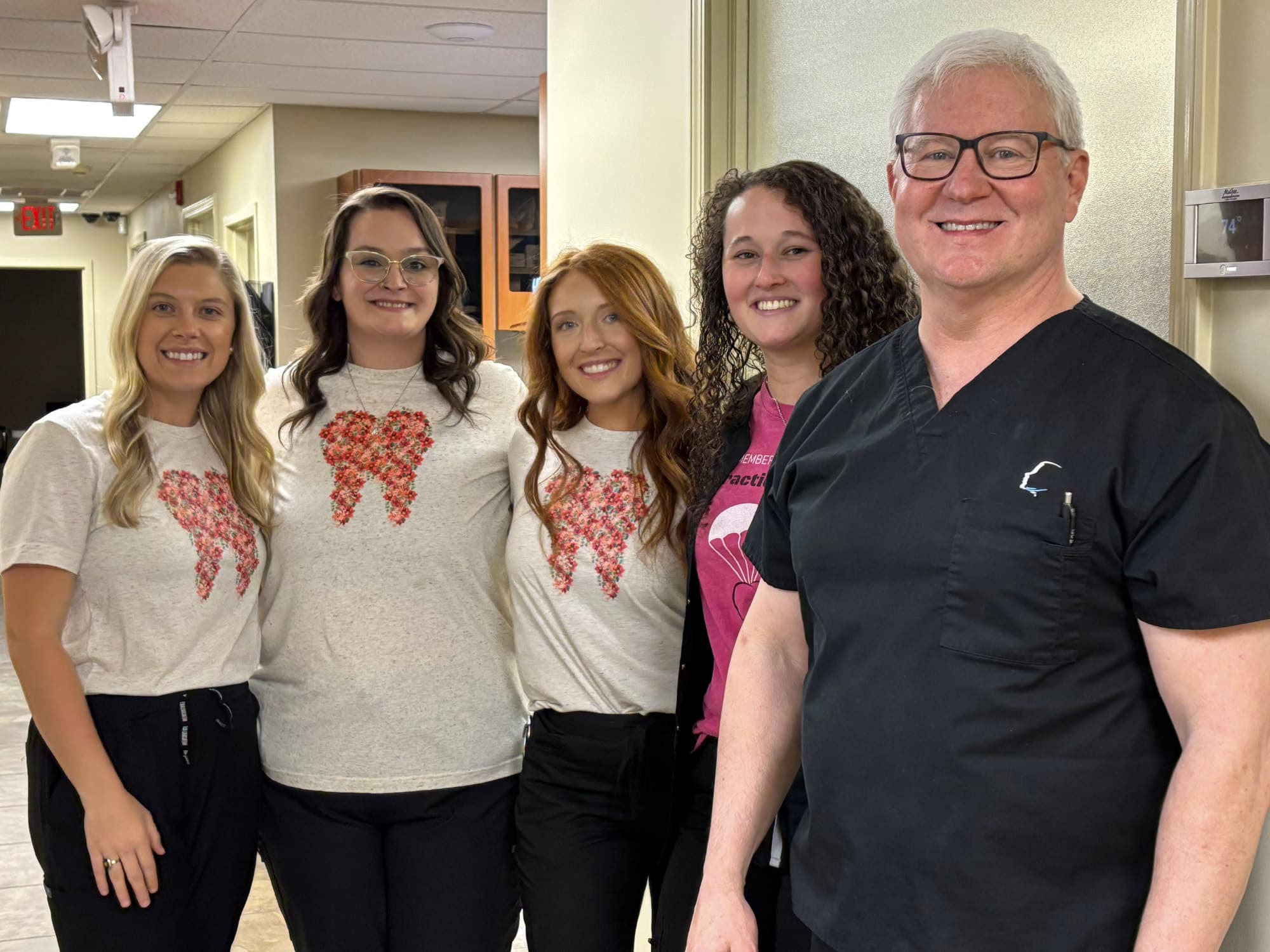 Five people smiling, standing indoors. Four wear tooth-themed shirts and one in scrubs. Professional office setting with visible ceiling tiles and lights.