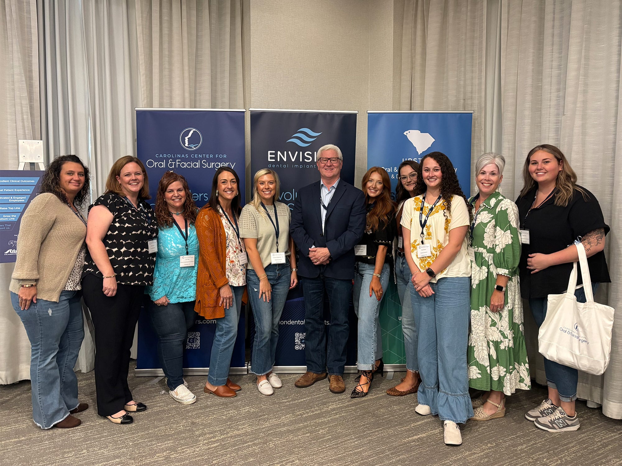 A group of people stand together, smiling, in front of banners for Carolinas Center for Oral & Facial Surgery and Envision Dental Implants.