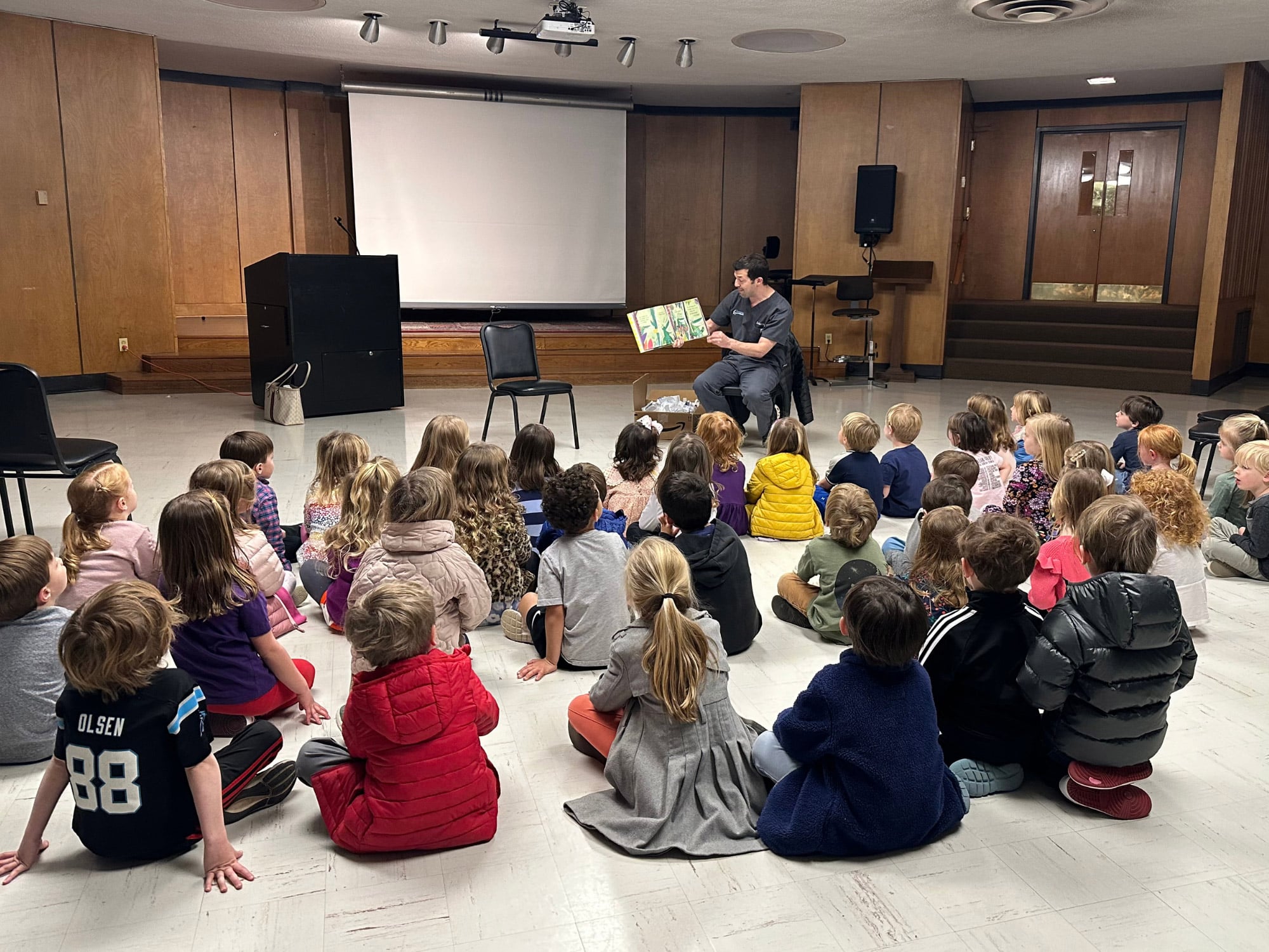 A group of children sits on the floor, listening to a person read a book in a classroom setting.