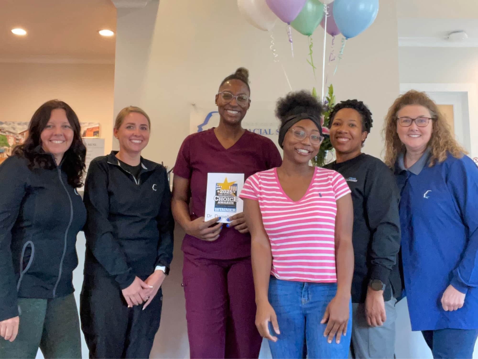 Seven people smiling indoors, one holding an award. They're gathered together with balloons, celebrating an achievement in a welcoming room setting.