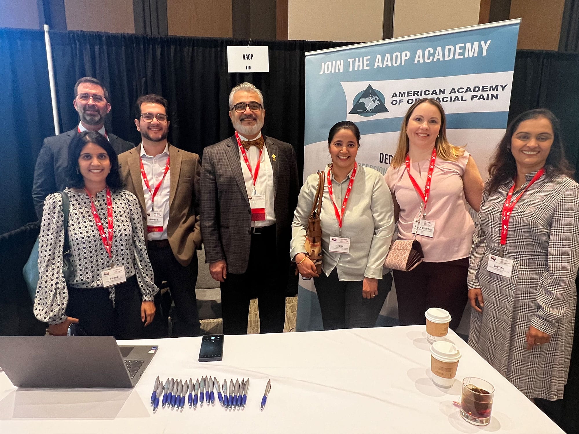 A group of seven people stands at an AAOP Academy booth. They are smiling and wearing name tags with red lanyards.