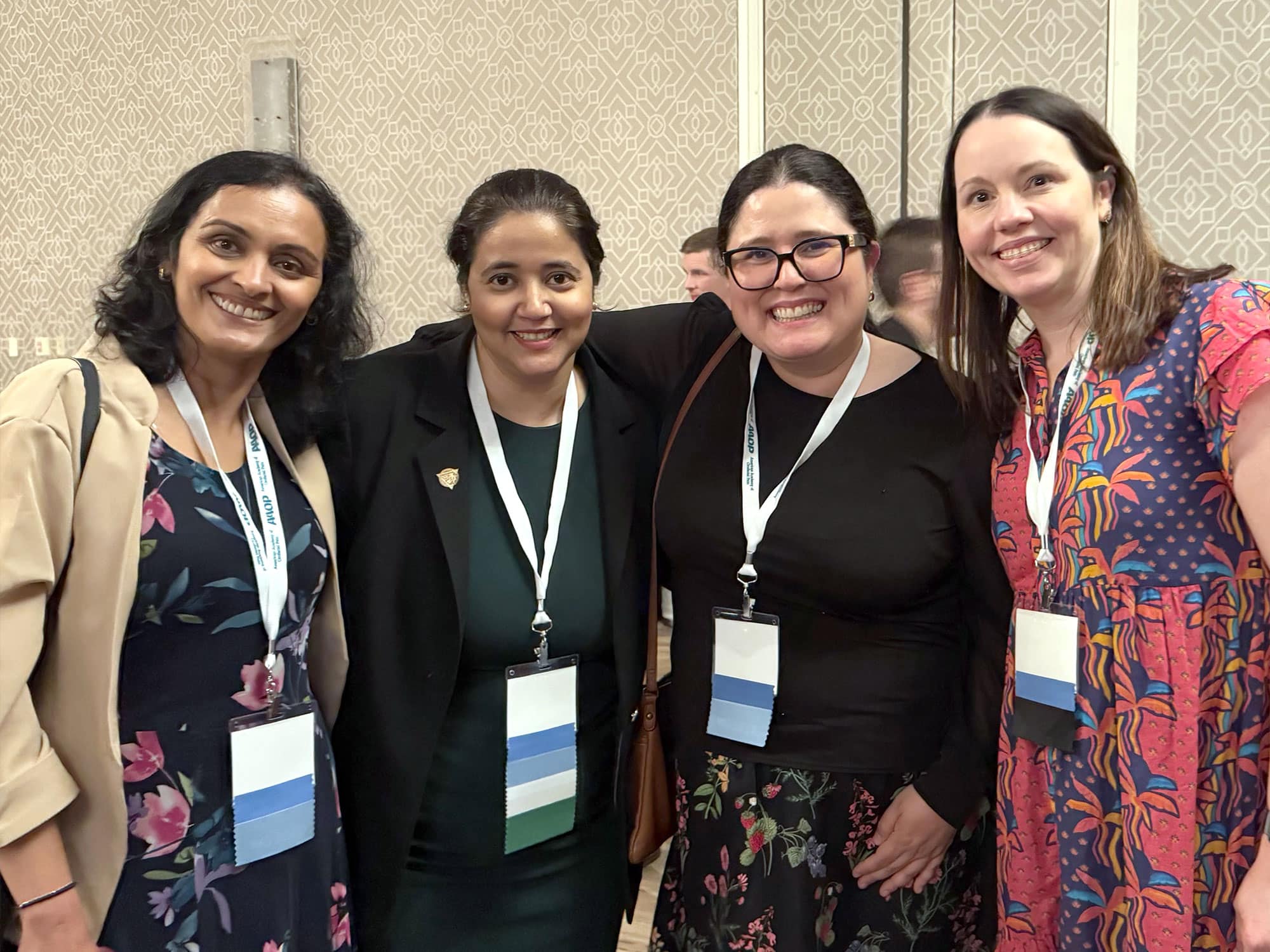 Four people smiling, wearing conference lanyards and badges, standing indoors against a patterned wall, suggesting a professional or networking event.