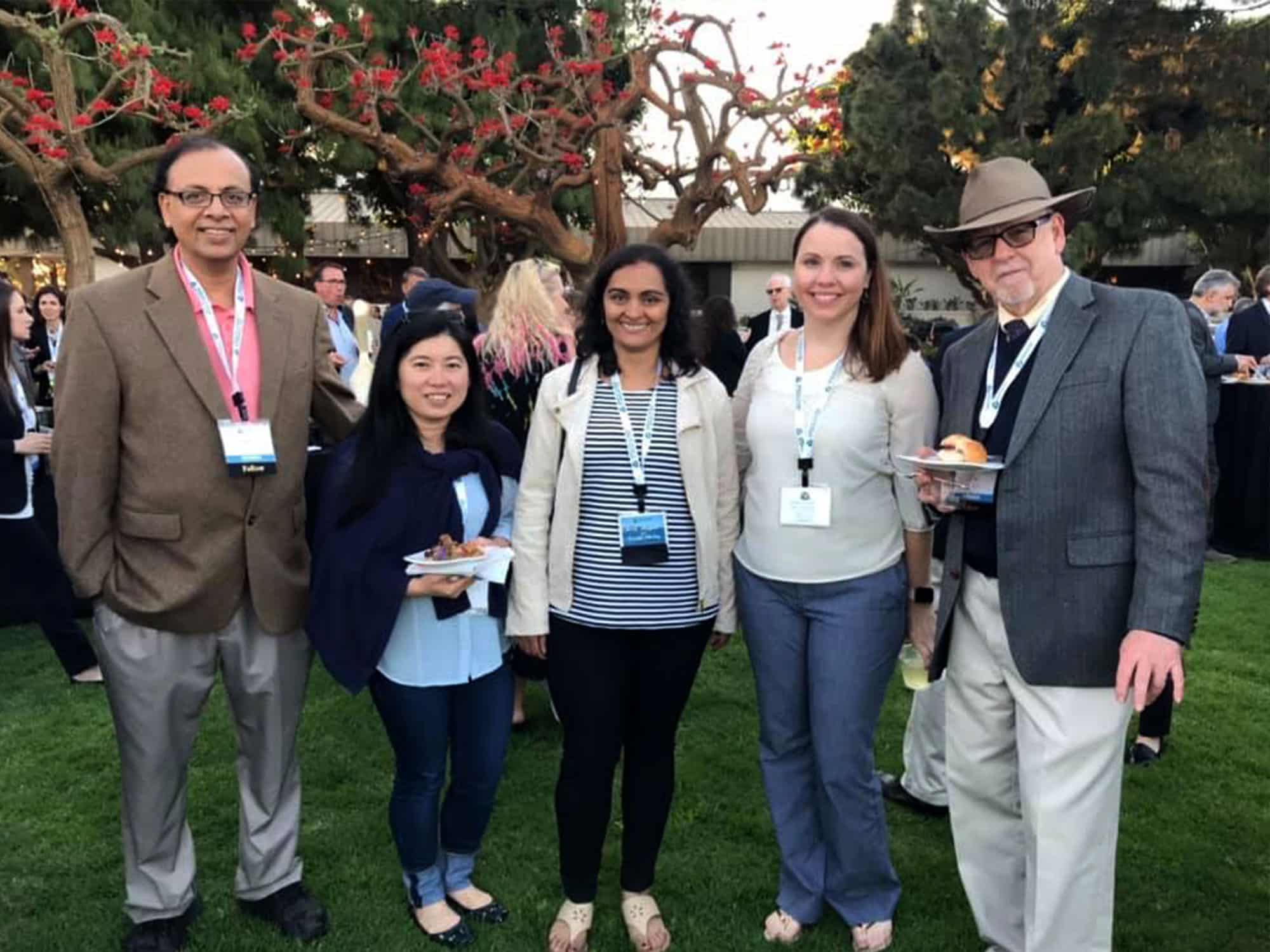 Five people with nametags, outdoors at a casual event, stand smiling near trees with red flowers, in a grassy area surrounded by others.