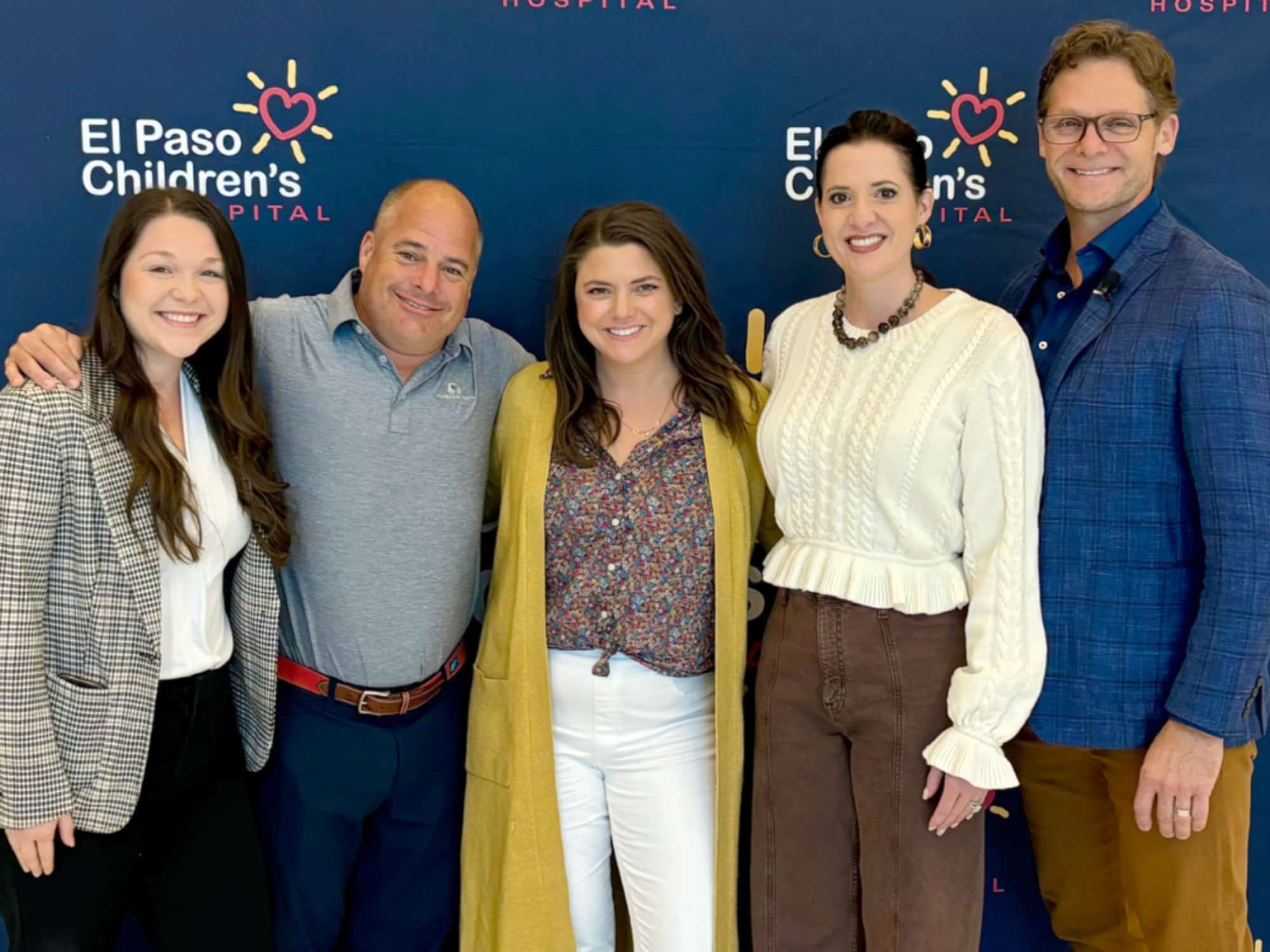 Five individuals smiling in front of an El Paso Children's Hospital backdrop, posing together.