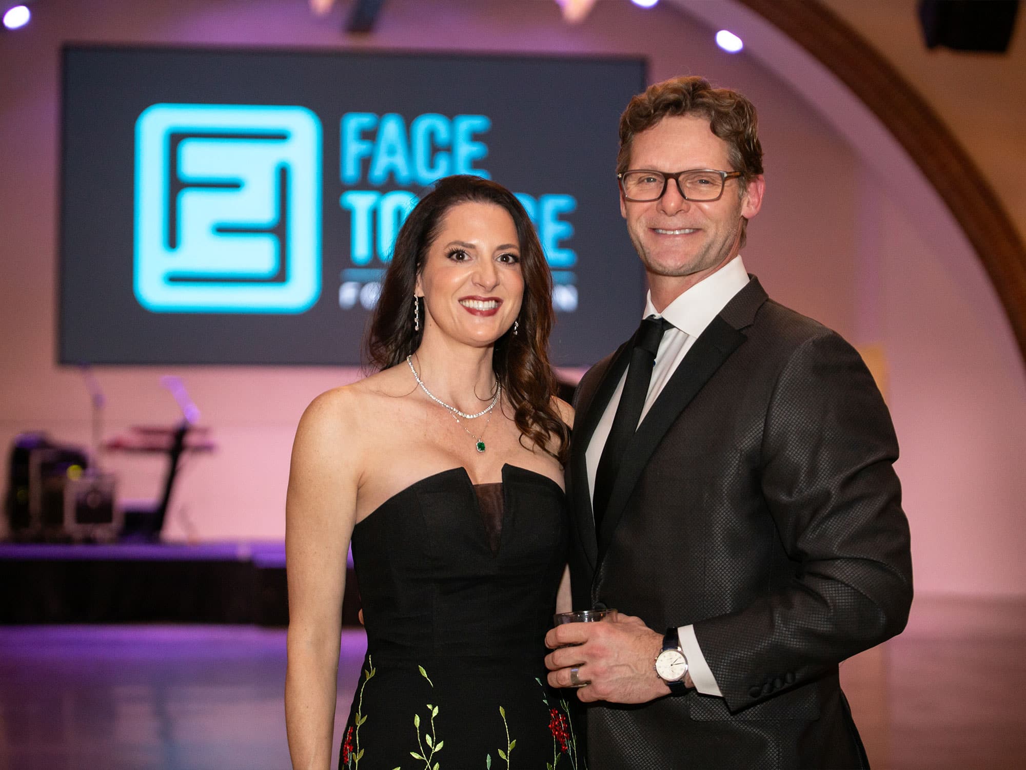 Two people in formal attire smiling, standing indoors with "FACE TO FACE" displayed on a screen behind them. Event setting, elegant atmosphere.