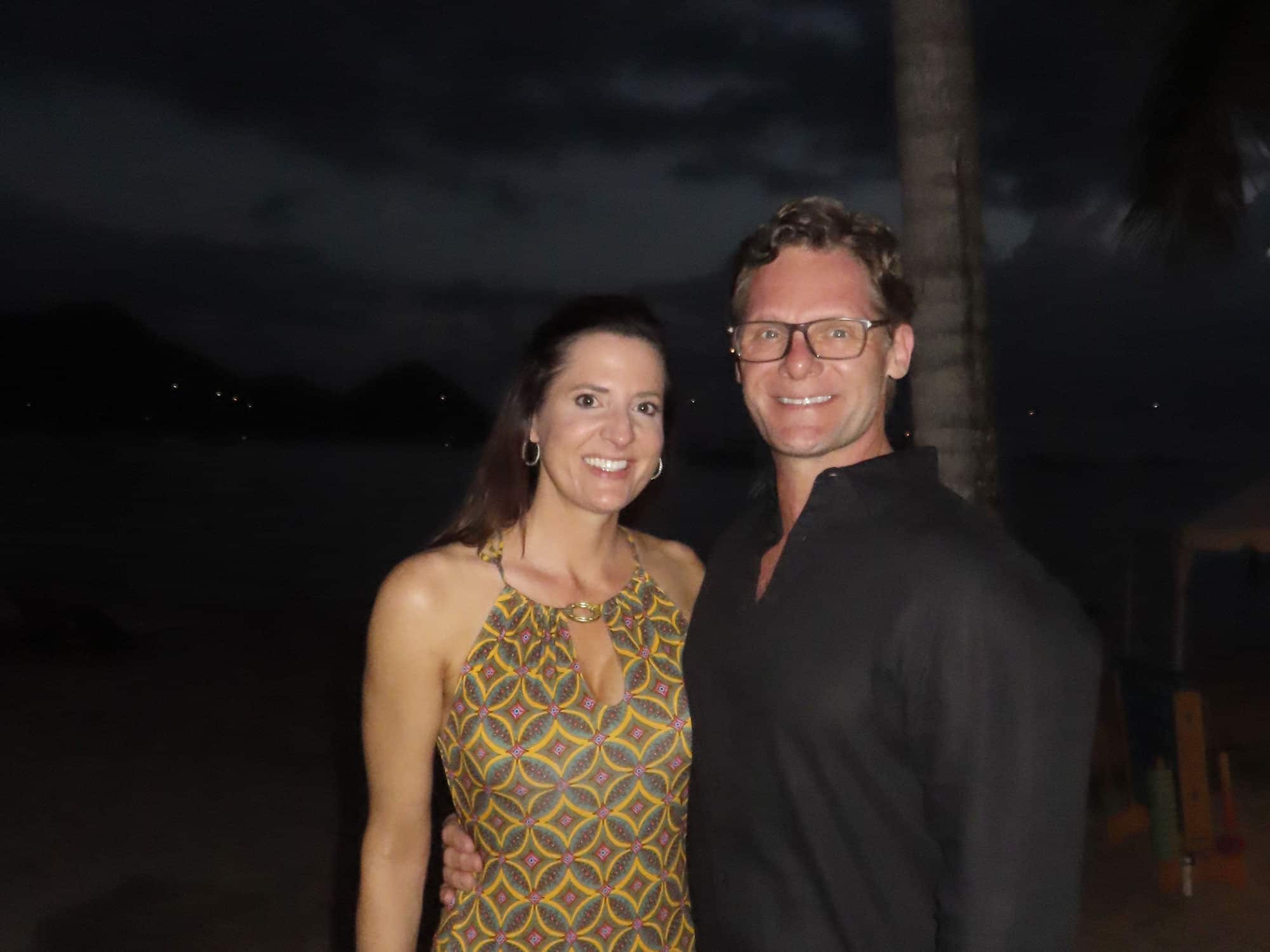 Two people smiling at night on a beach, with a palm tree and dark mountains in the background. No landmarks visible.