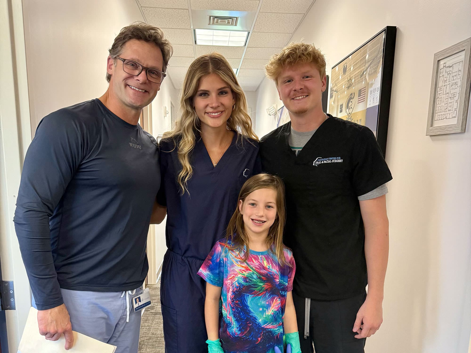 Three people in scrubs and a child smile in a hallway, standing together. A framed photograph is visible on the wall.