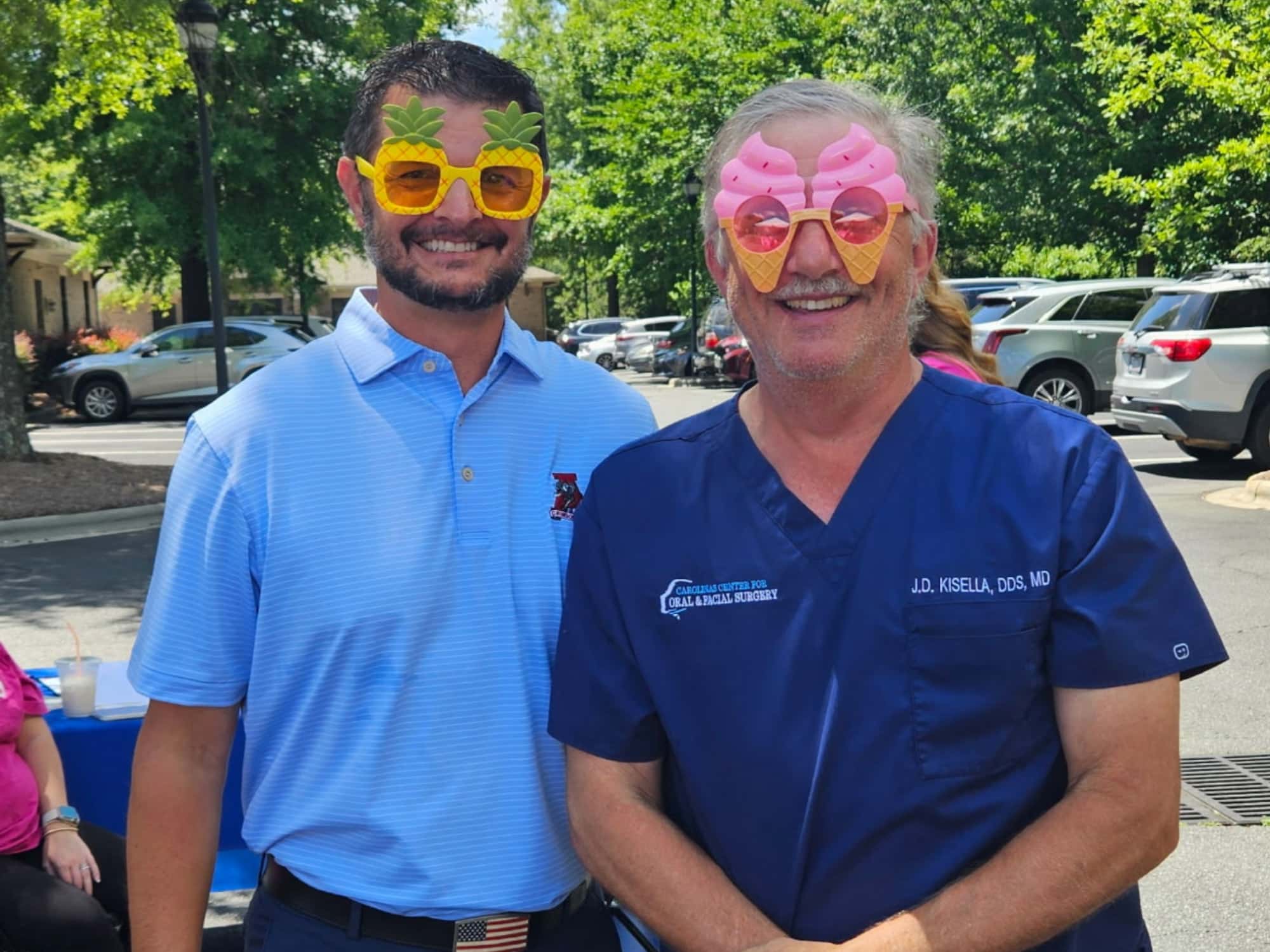 Two persons wearing playful sunglasses stand outdoors, smiling. One wears a blue polo; the other, blue scrubs. Trees and parked cars in the background.