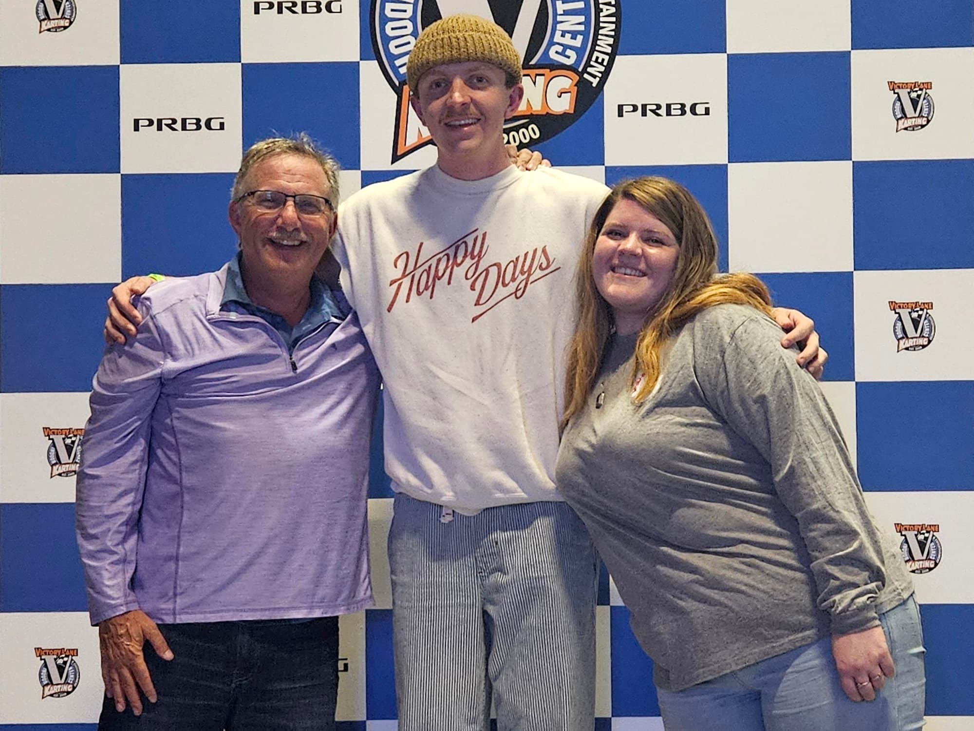 Three people standing together in front of a checkered wall featuring various logos, including "Victory Lane Karting."