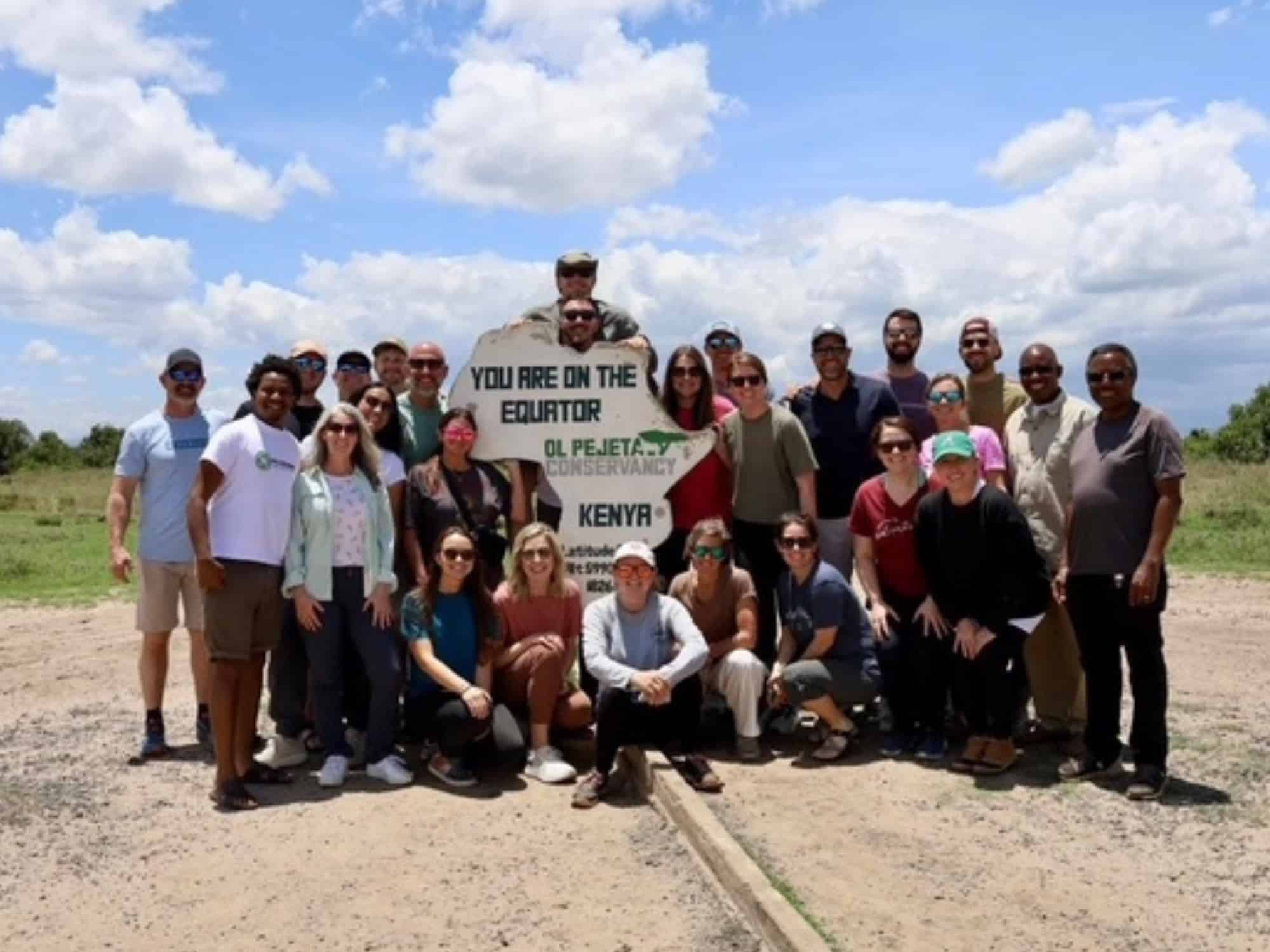 A group of people poses around an "Equator" sign in Ol Pejeta Conservancy, Kenya, on a sunny day, with a bright blue sky.