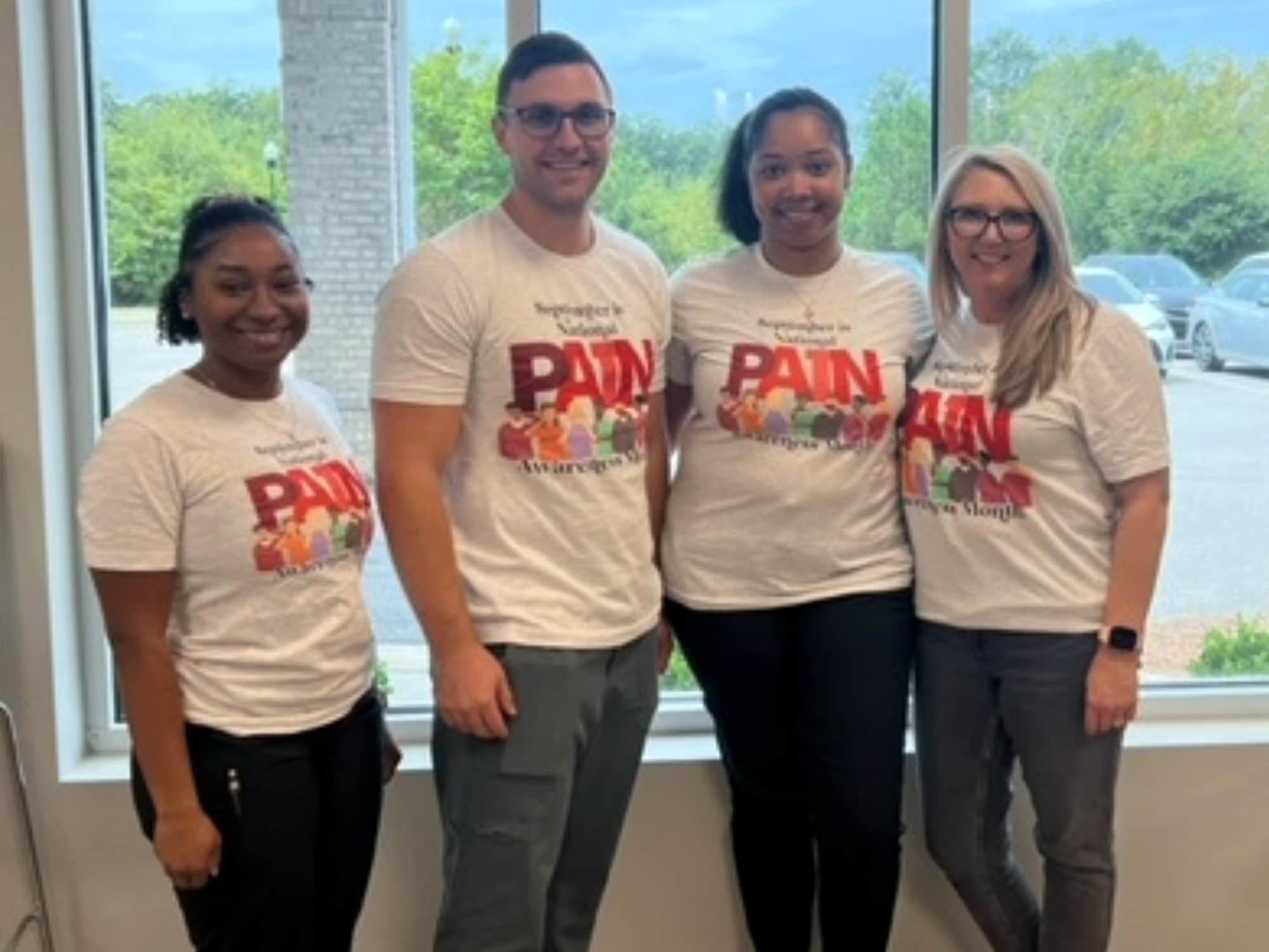 Four people wearing matching shirts stand indoors near a window, smiling. Trees and cars are visible outside. No landmarks or historical buildings present.