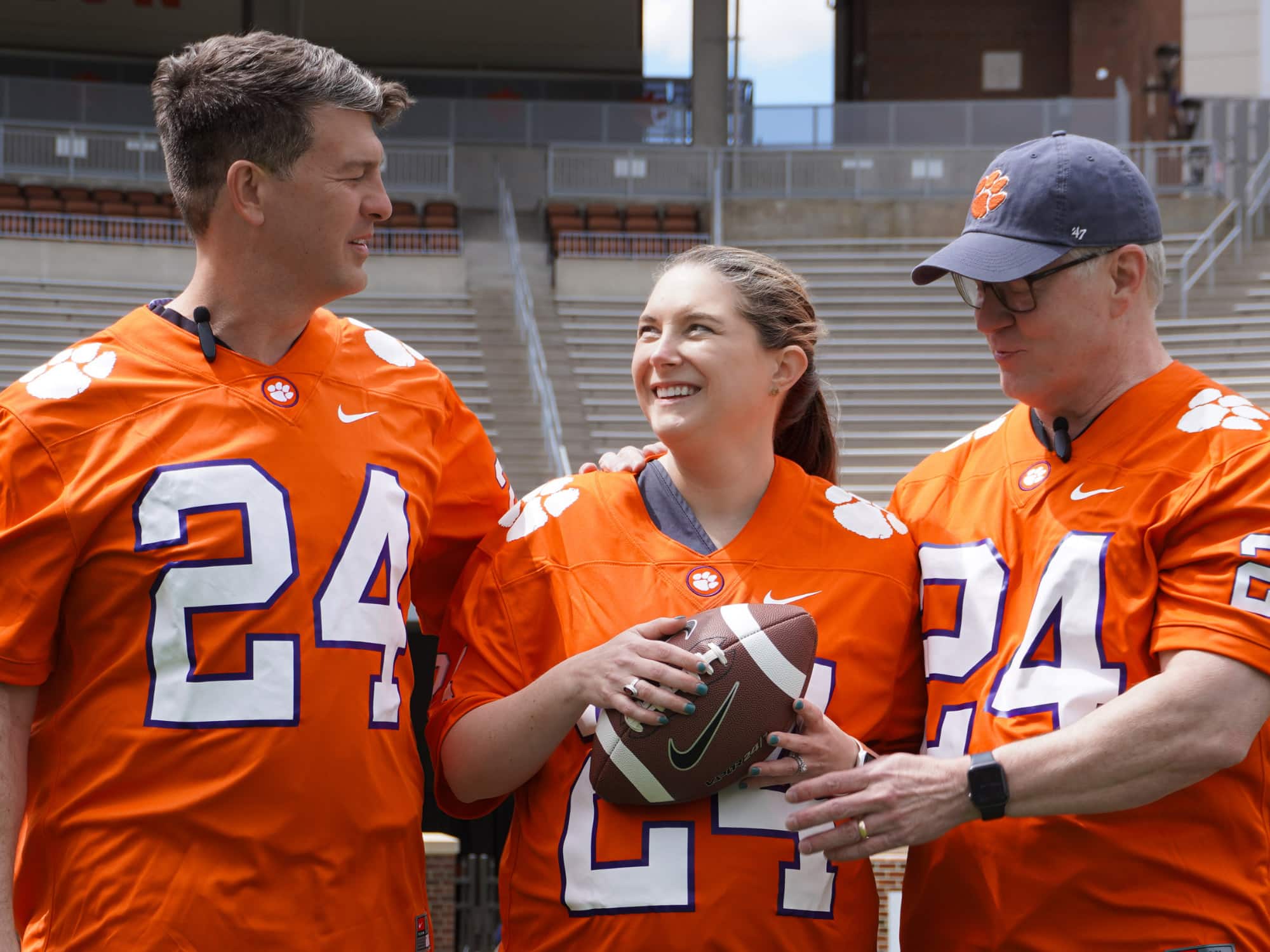 Three people wearing matching orange Clemson jerseys, standing in a stadium, smiling and holding a football.